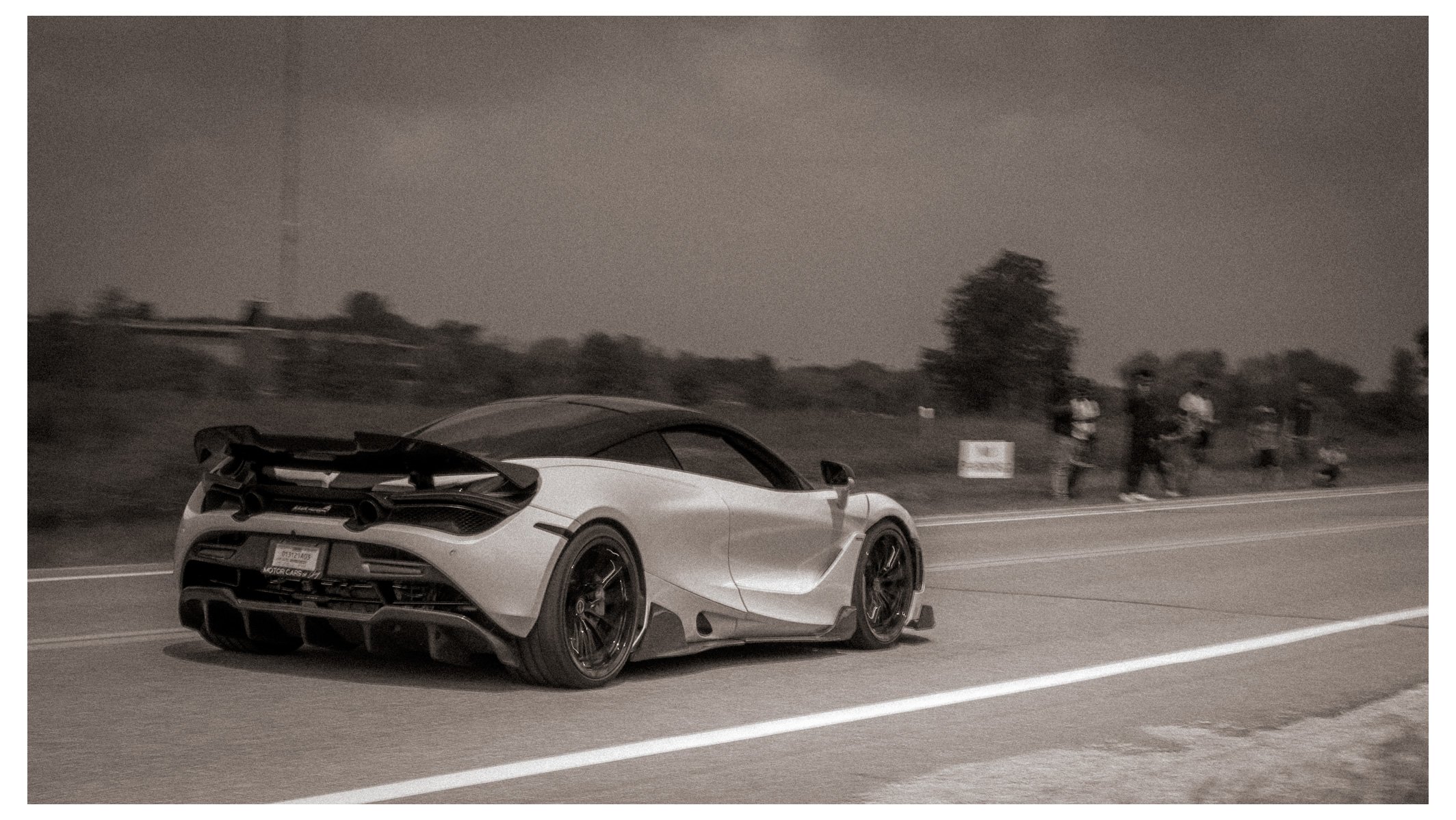 A white sports car with a black rear wing on a highway at night, with a line of trees and a group of people walking along the side of the road in the background.