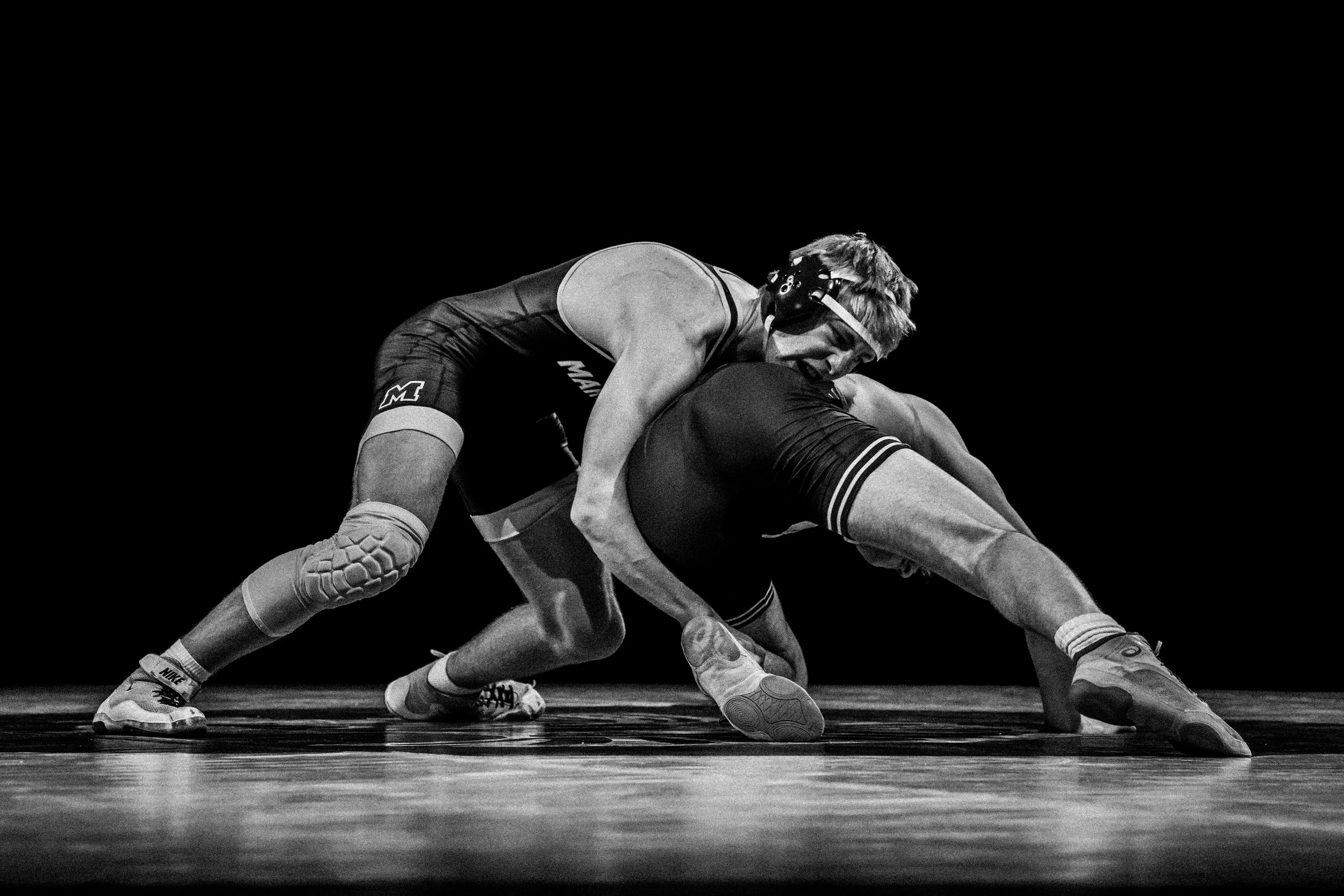Black and white photo of two wrestlers in a match on a wrestling mat. One wrestler is bent forward while the other is positioned over him, both wearing singlets and wrestling shoes.