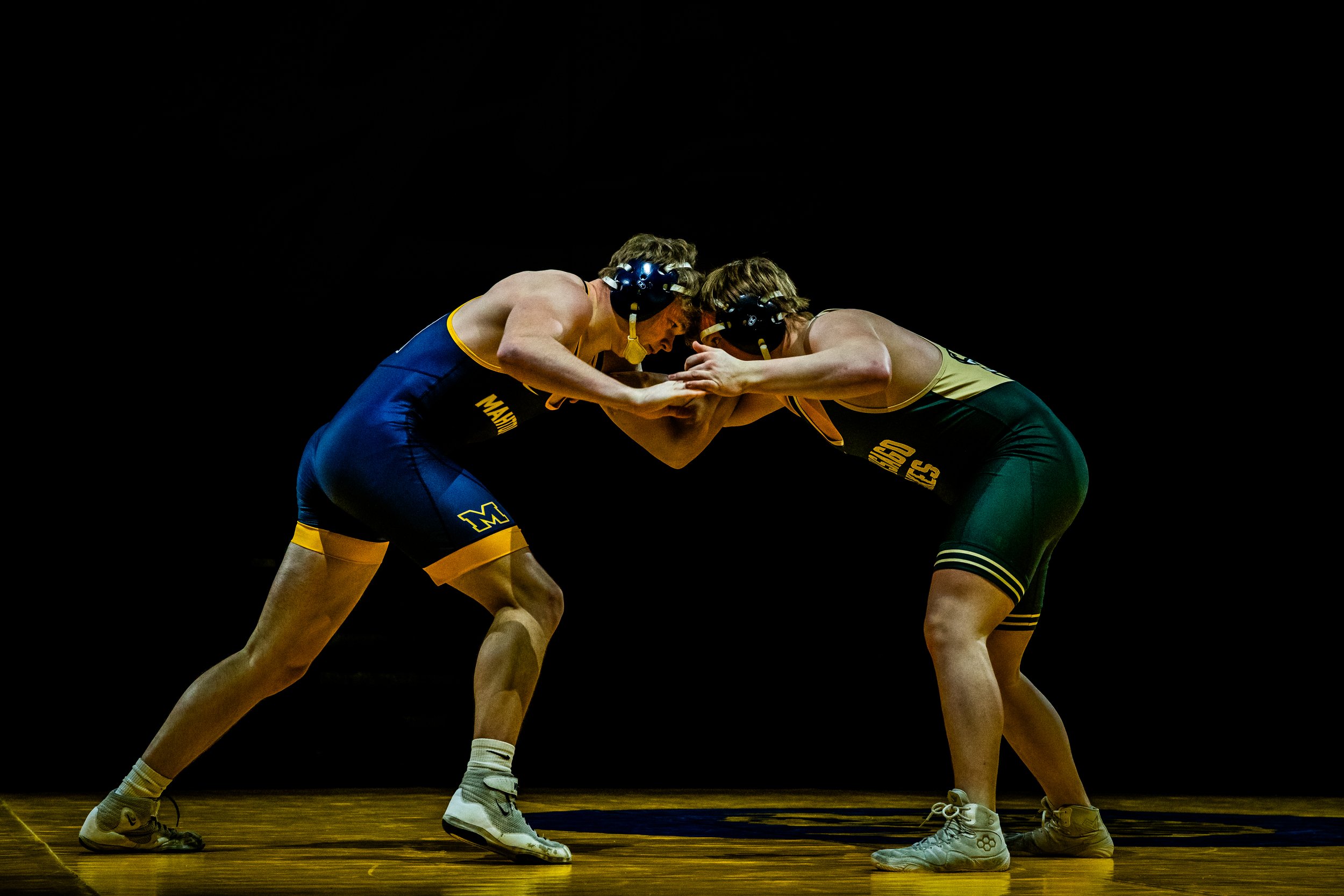 Two male wrestlers in a match, wearing singlets and headgear, engaged in a grapple on a wooden wrestling mat against a black background.