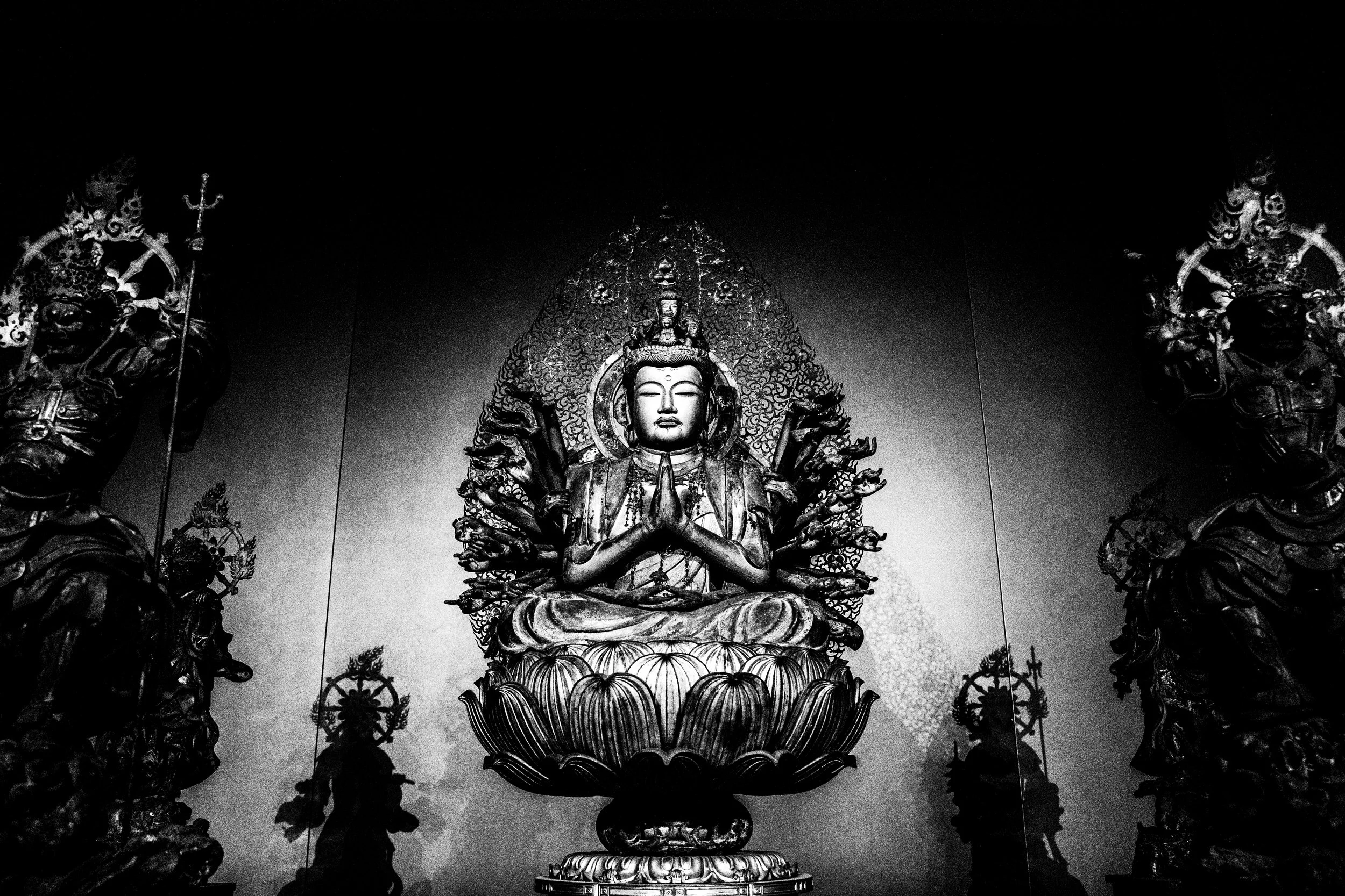 Black and white image of a large, seated Buddhist figure with multiple arms, surrounded by ornate statues of warriors on either side.