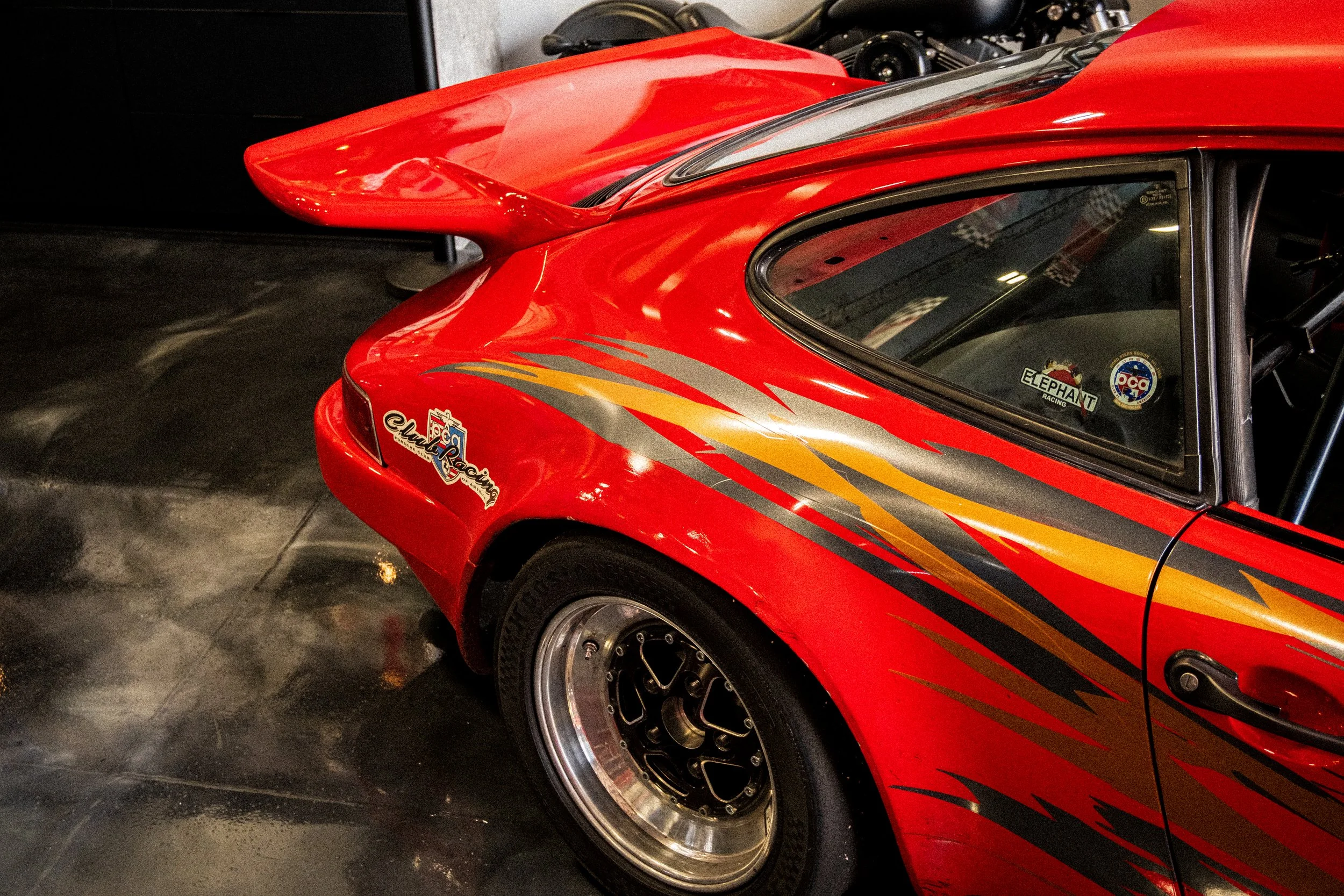 Close-up of a red race car with custom decals and wide tires, showing part of the front and side, on a dark tiled floor.