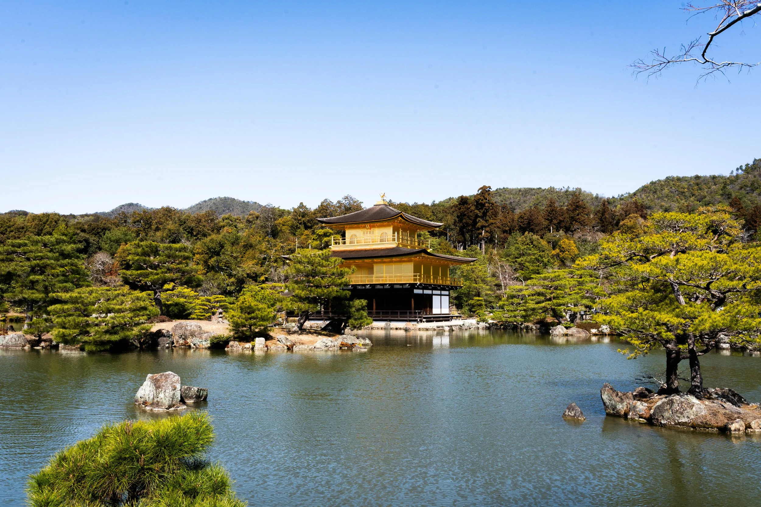 A traditional Japanese building with a pagoda-style roof surrounded by lush greenery, trees, and water under a clear blue sky.