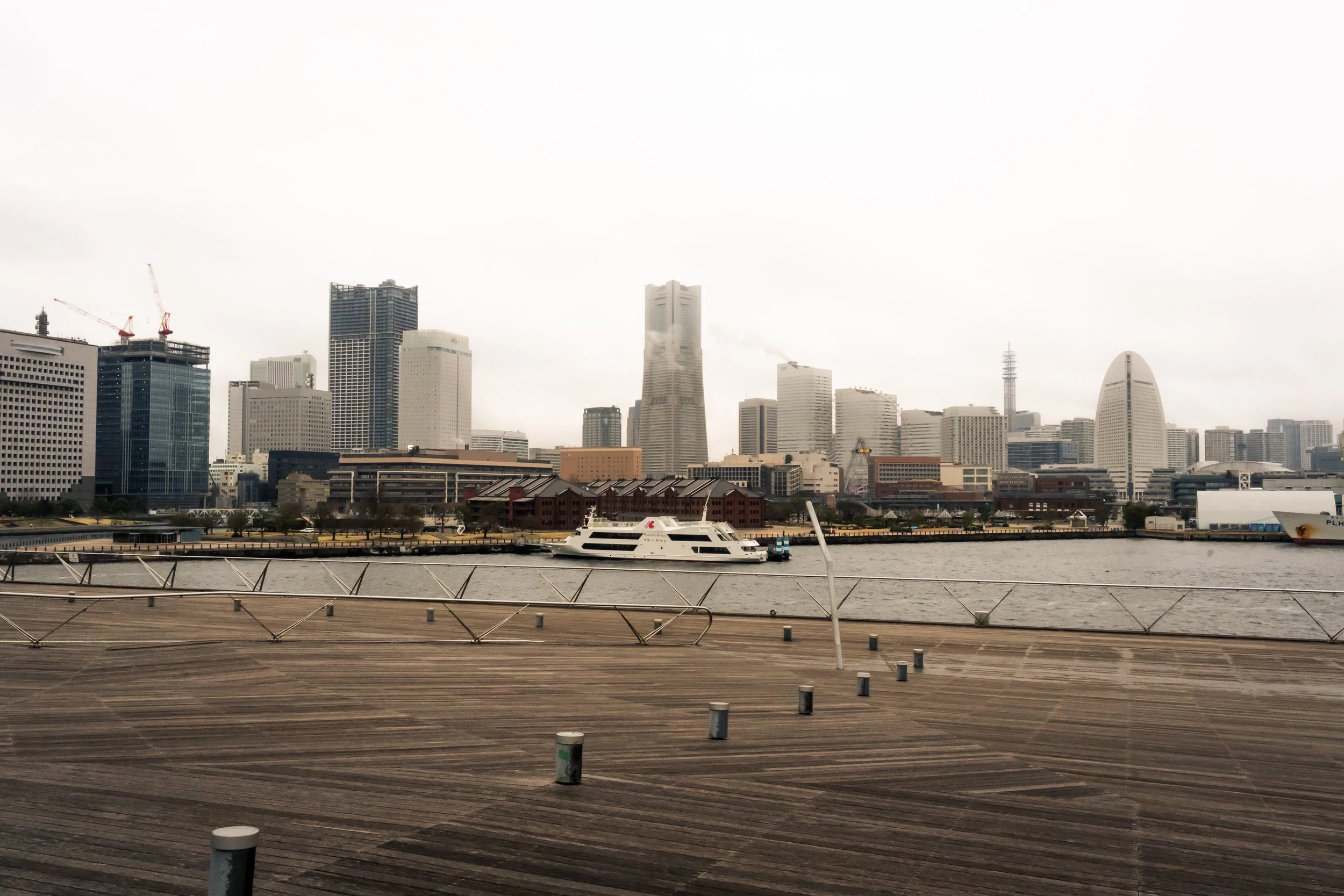 City skyline with tall buildings, a waterfront, and a boat docked along the pier, overcast sky.