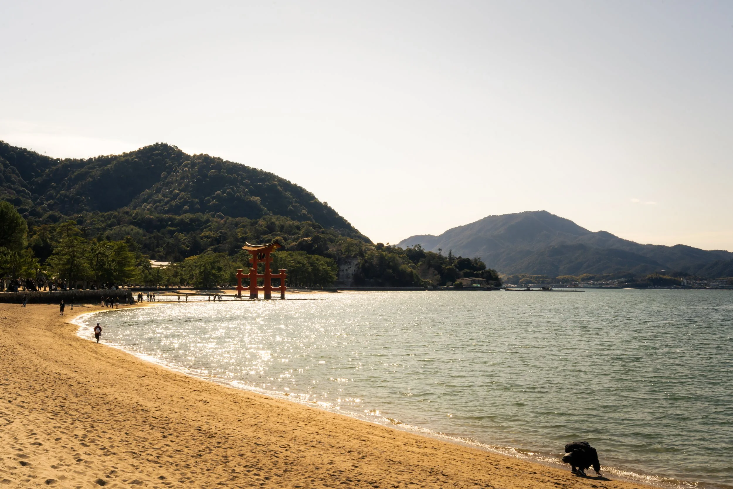 A scenic view of a beach with golden sand, gentle water, and lush green mountains in the background. A small red torii gate stands near the shoreline, and a few people are walking along the beach and in the water.