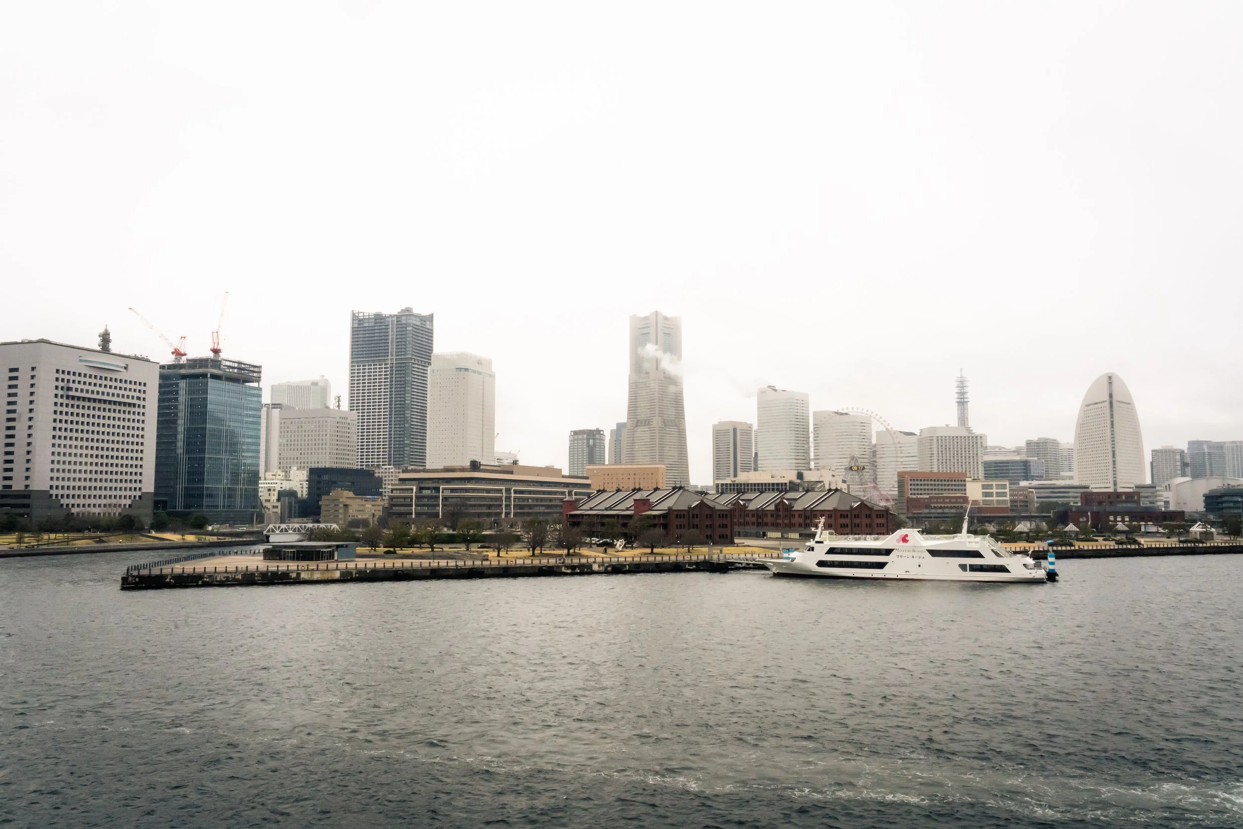 A city skyline with tall buildings, some under construction, seen across a body of water with a yacht docked at a pier in the foreground.