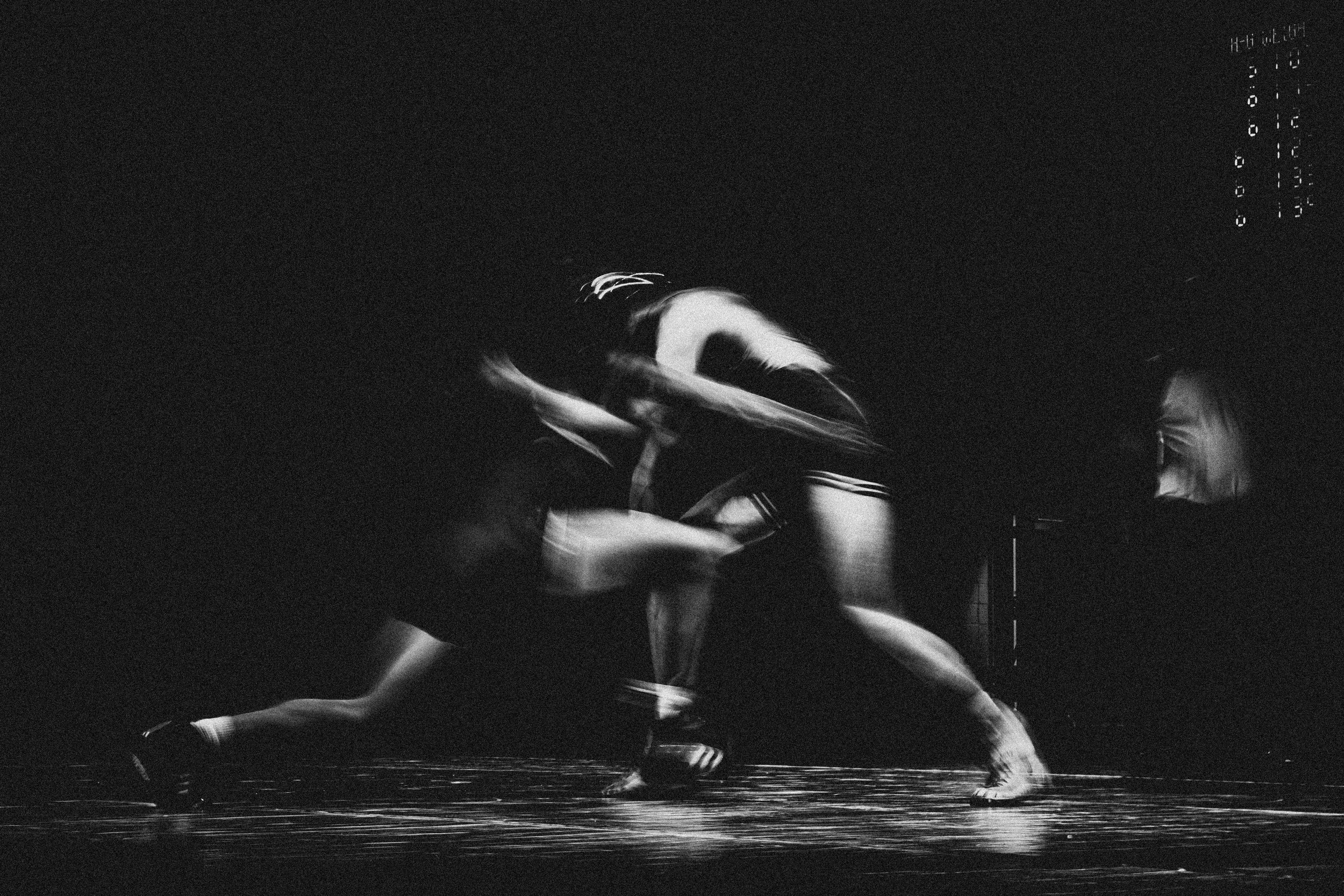 A black and white photograph of a couple dancing closely, with their bodies intertwined and hands holding each other, set against a dark background with reflections on the floor.