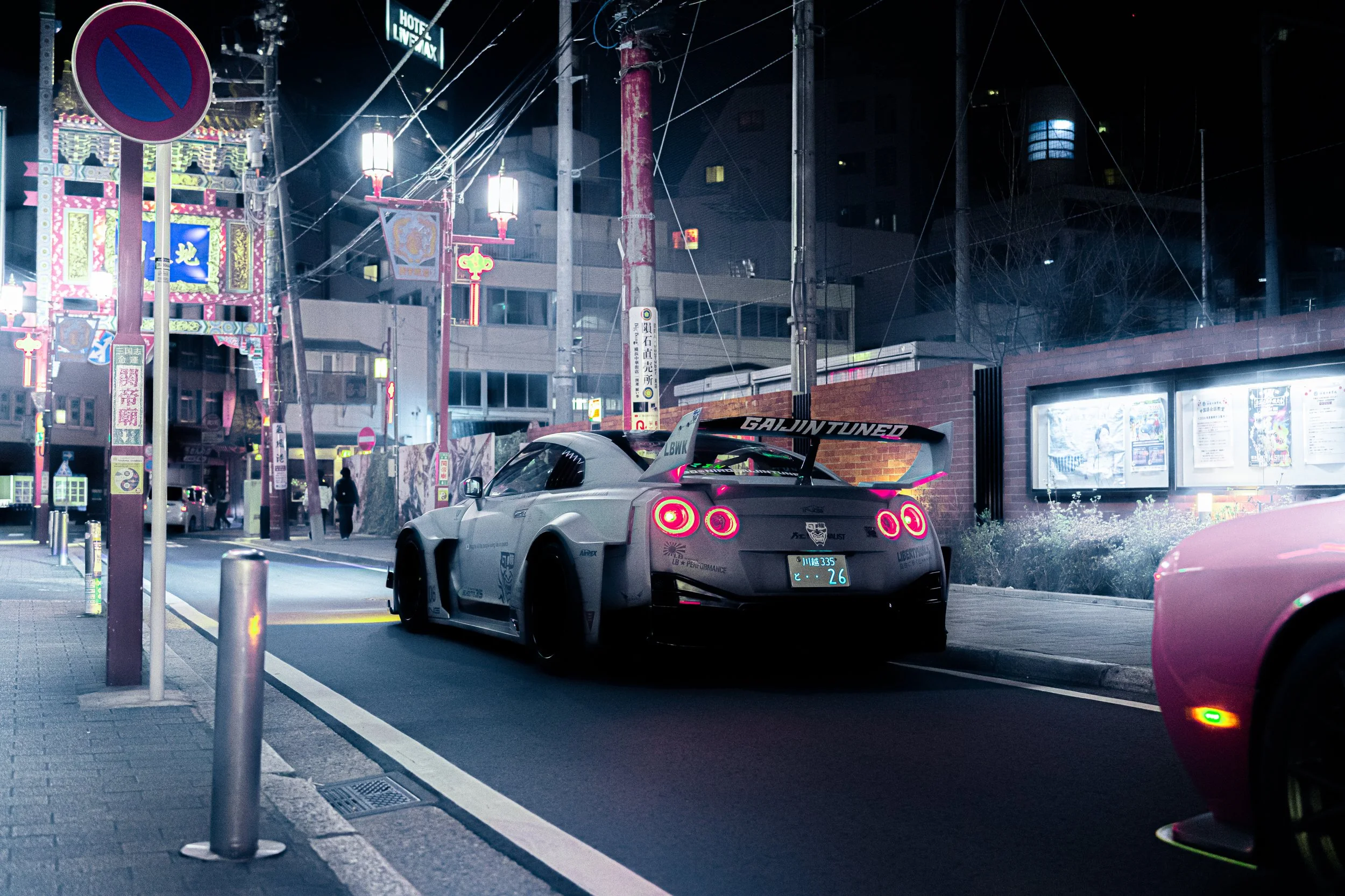 Nighttime urban scene with a silver sports car parked on the street, illuminated signs and buildings in the background, and a no parking sign on the sidewalk.