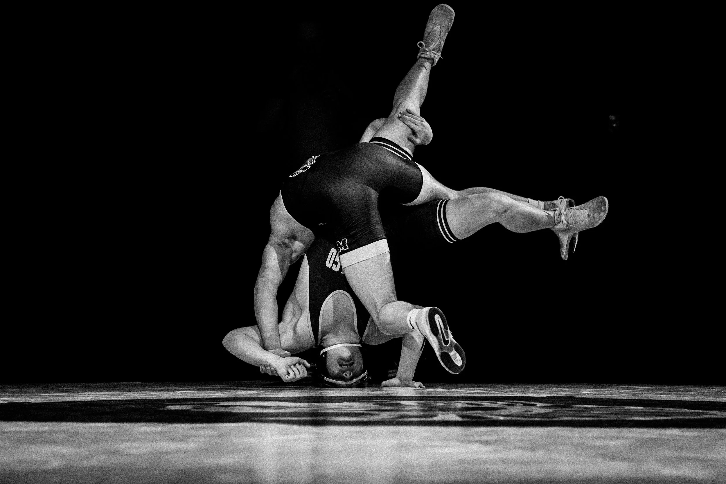 Black and white photograph of two wrestlers in a match, one in the process of pinning the other to the mat. The wrestler on top is holding the opponent's head and arm, with a partially visible face showing concentration. The wrestler on the bottom is