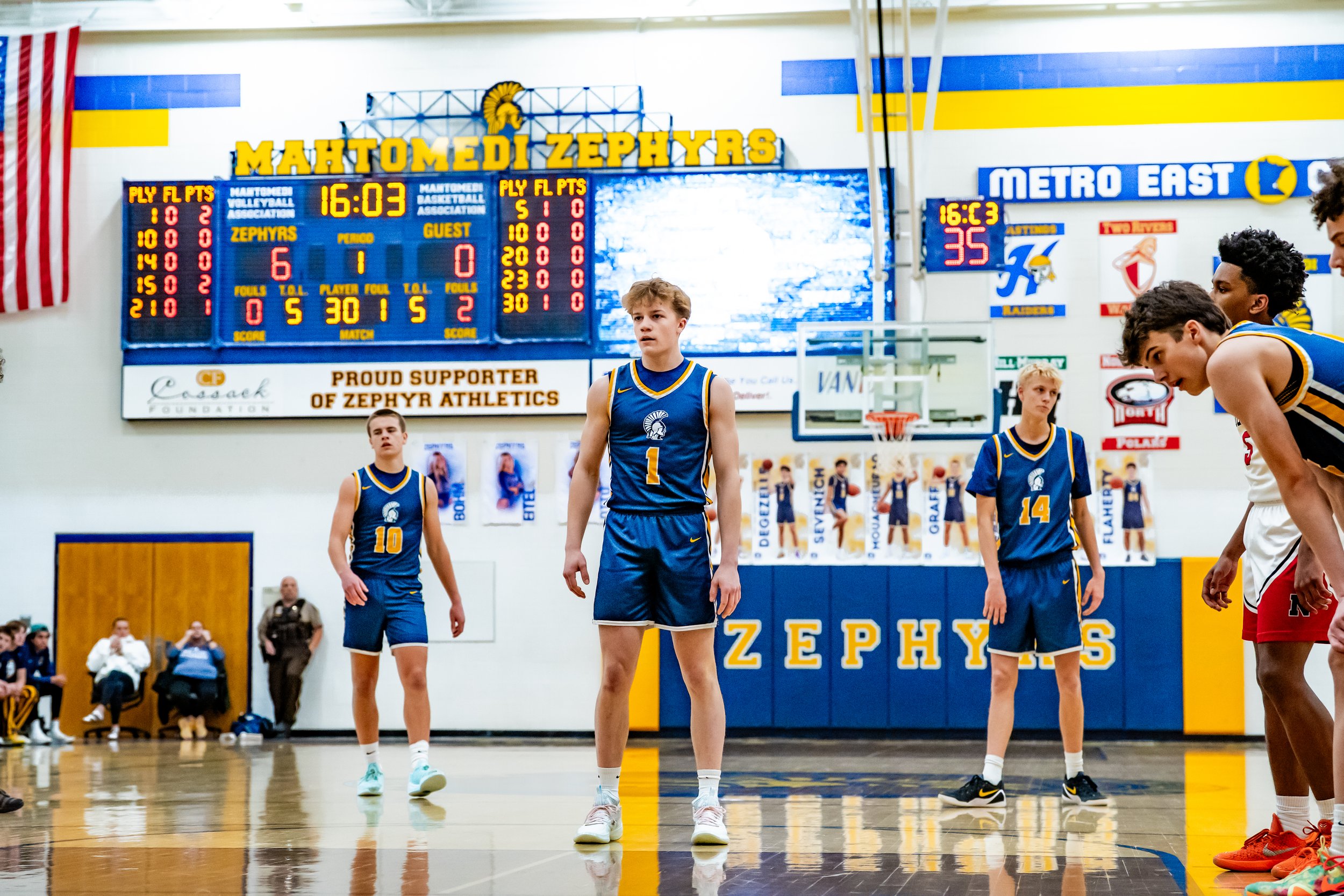 Young basketball players standing on a court during a game at Mahtomediz Zephyrs gym, with scoreboard and banners visible.