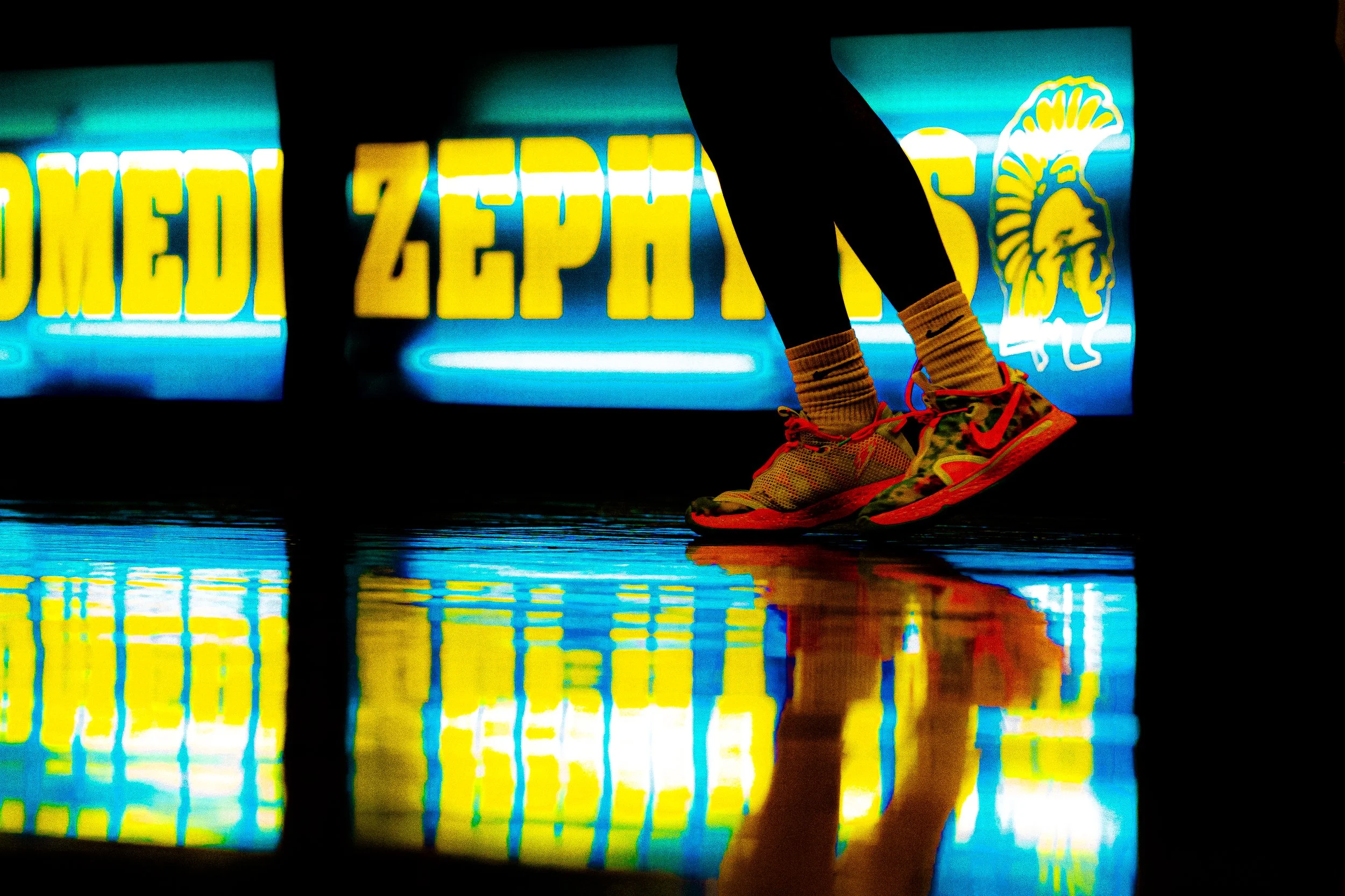 Person wearing colorful running shoes and black leggings, standing on a reflective floor with neon-yellow and blue sign in the background.