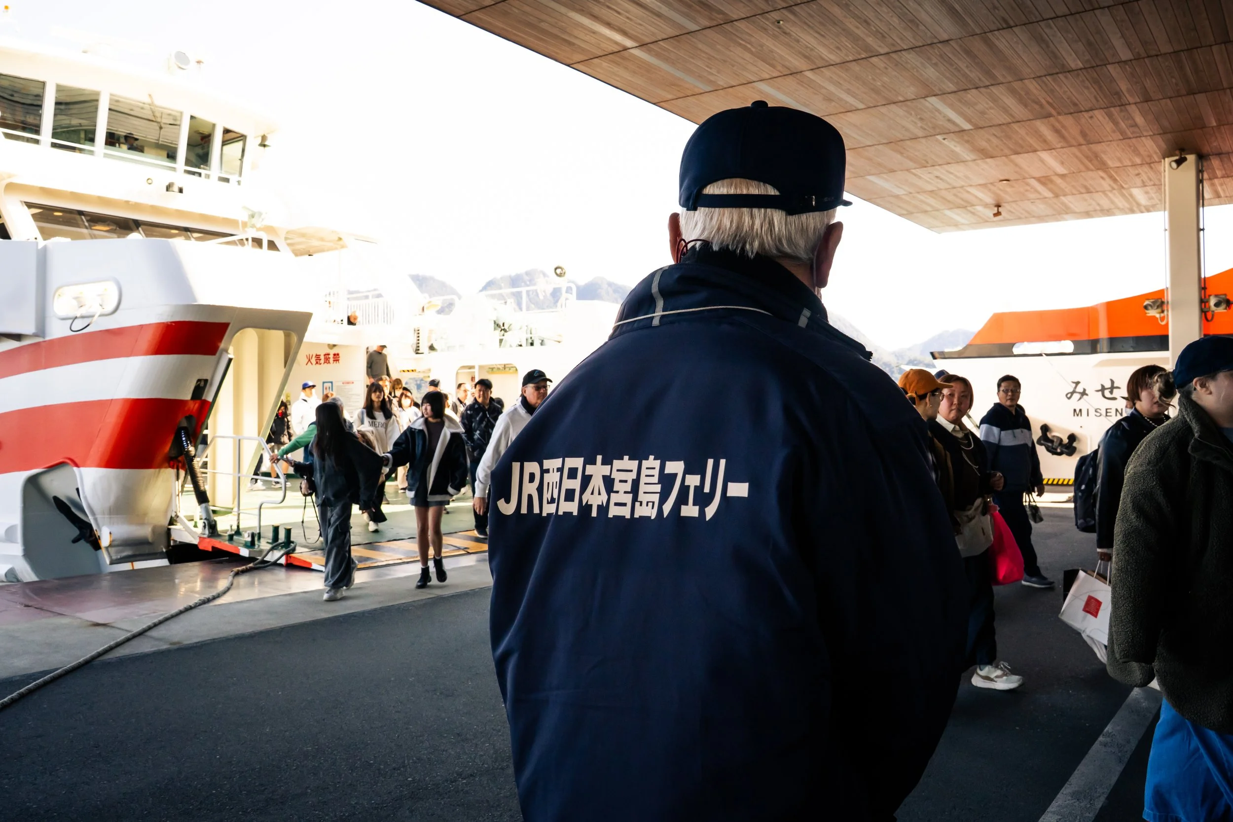 A man with gray hair wearing a black cap and a navy jacket with Japanese writing on the back is standing at a ferry terminal. In the background, there are many people waiting, boats docked, and a building with large glass windows, suggesting a busy p