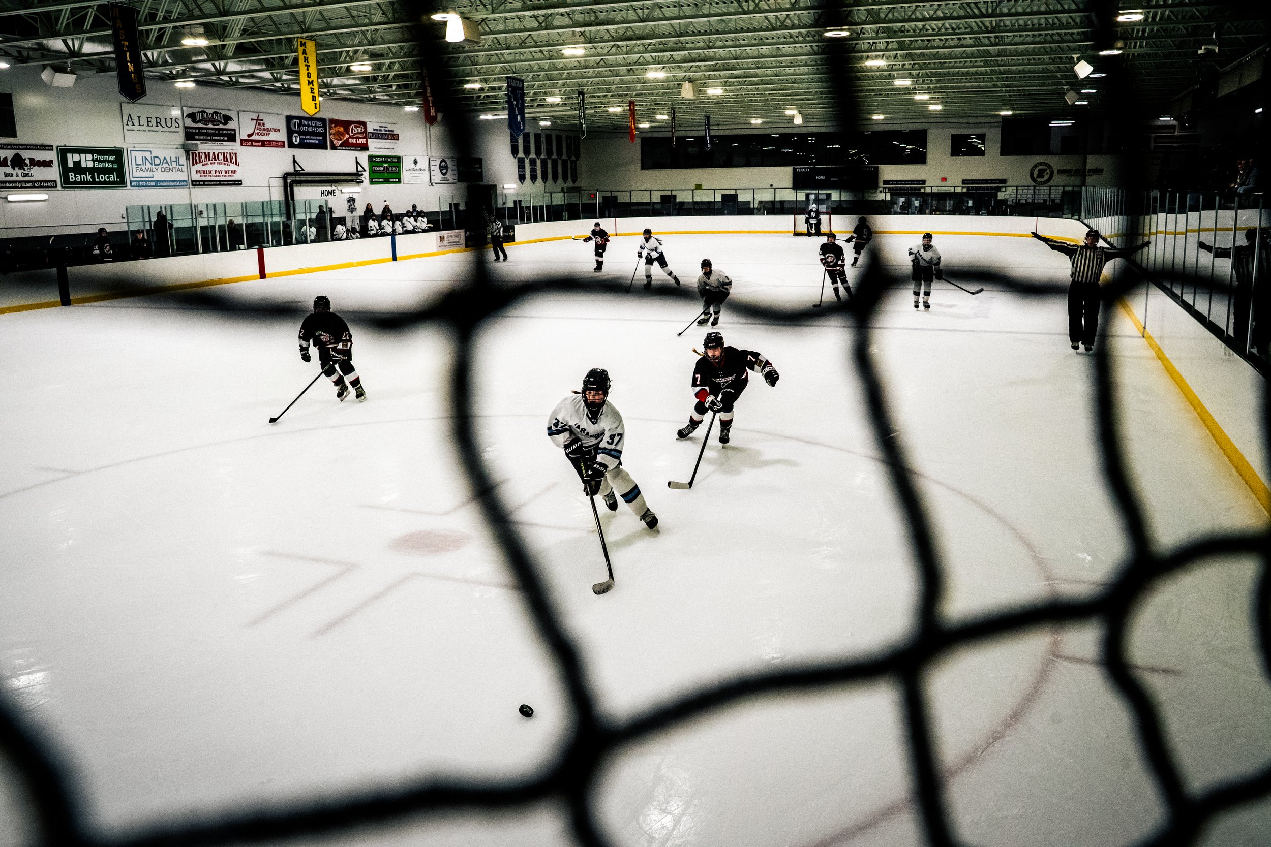 Ice hockey game at an indoor rink with players on the ice and spectators on the bench, viewed through a chain-link fence.