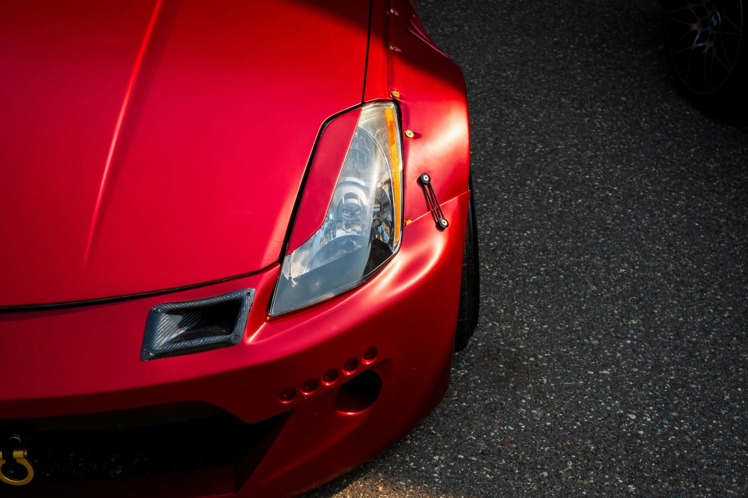 Close-up of the front of a red car, showing part of the headlight, hood, and a carbon fiber vent on the side. The car is parked on a dark, textured asphalt surface.