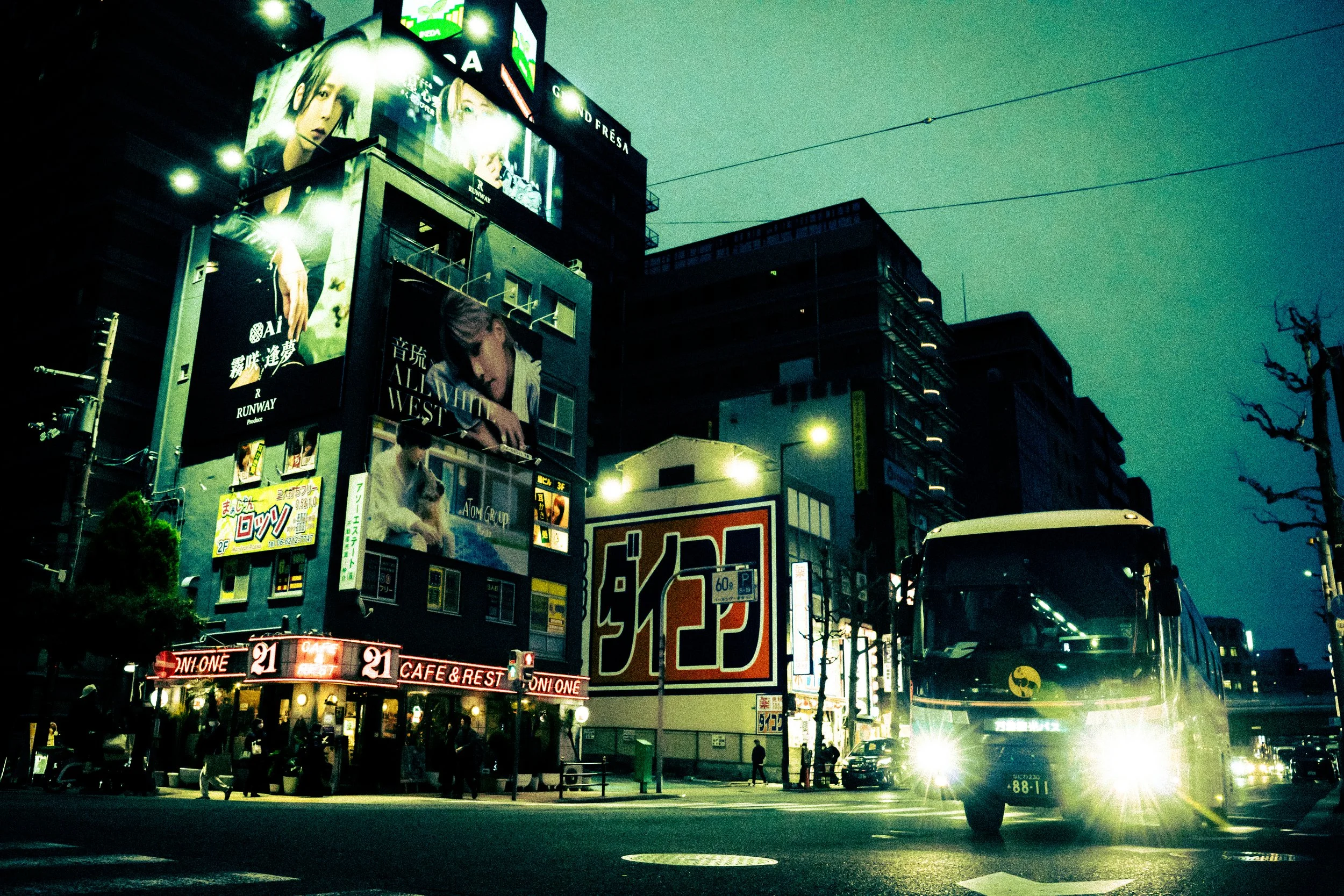 A city street at night with illuminated billboards and signs on buildings. There are several advertisements, some featuring models, and a street filled with cars and pedestrians. Vehicles' headlights are on, and the scene is lively despite the darkne