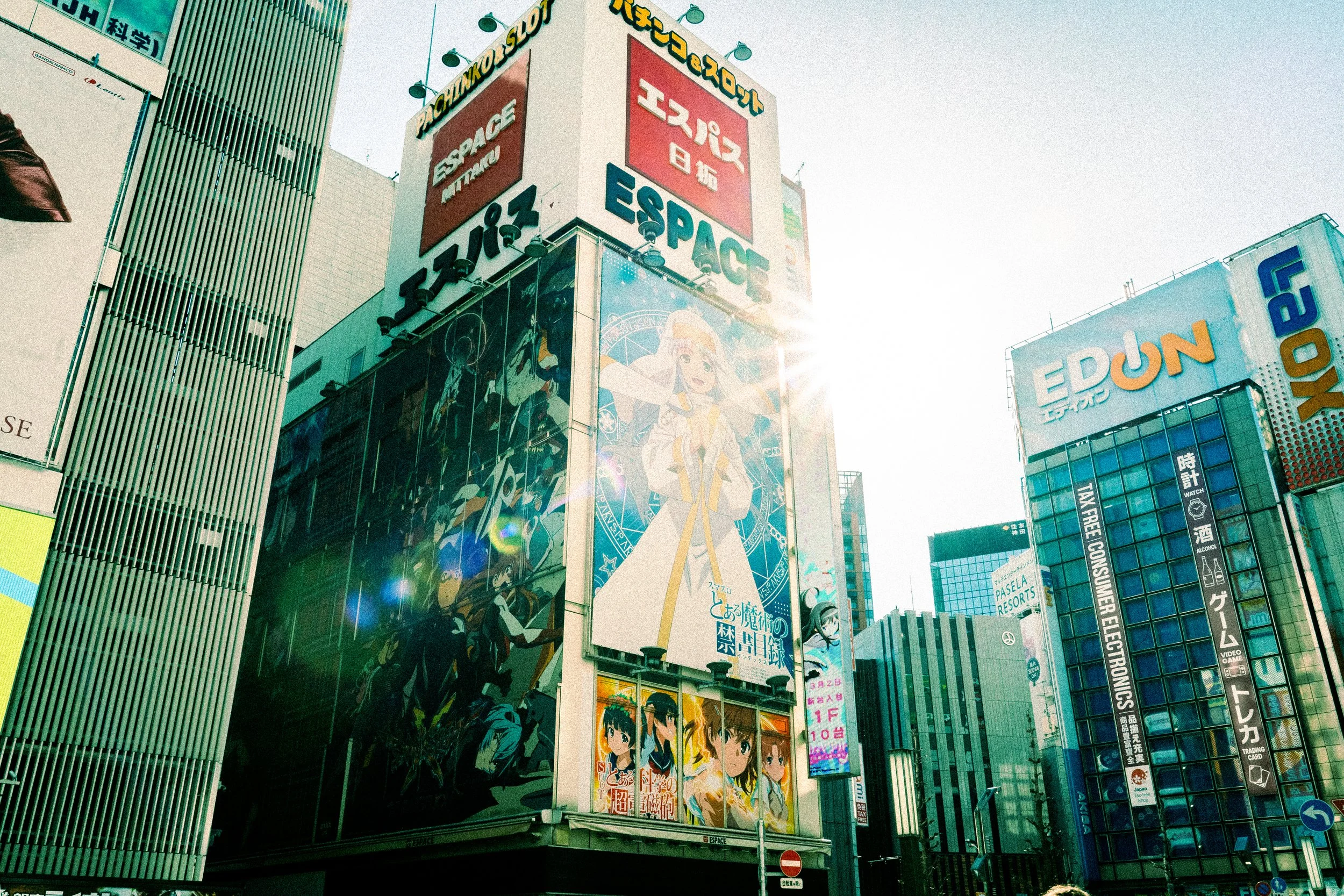 Billboards and electronic signs in a busy city, featuring anime and manga style characters, with some signs in Japanese and English, under a clear sky.