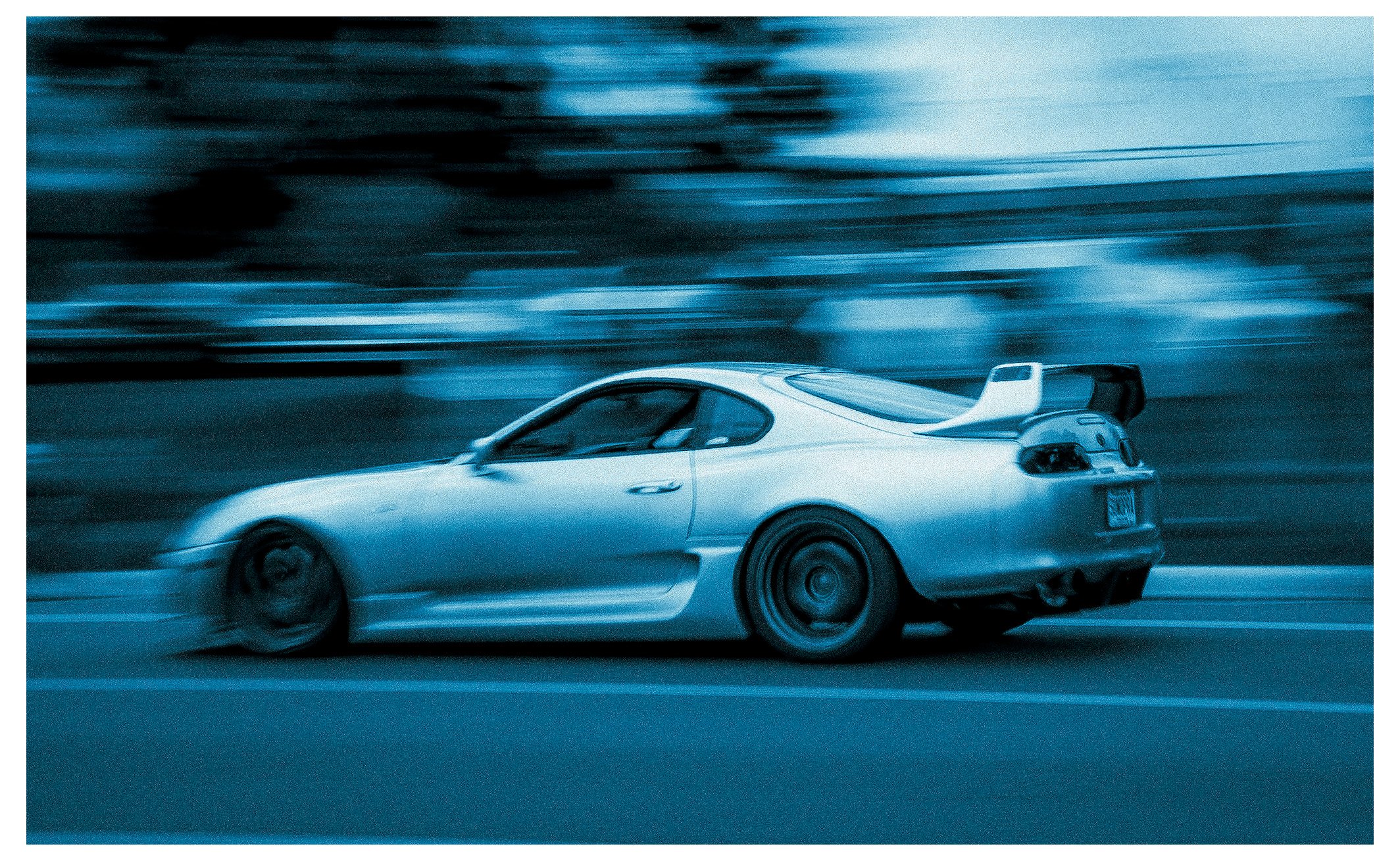 A silver sports car driving fast on a road, blurred background indicating motion, during twilight or dusk.