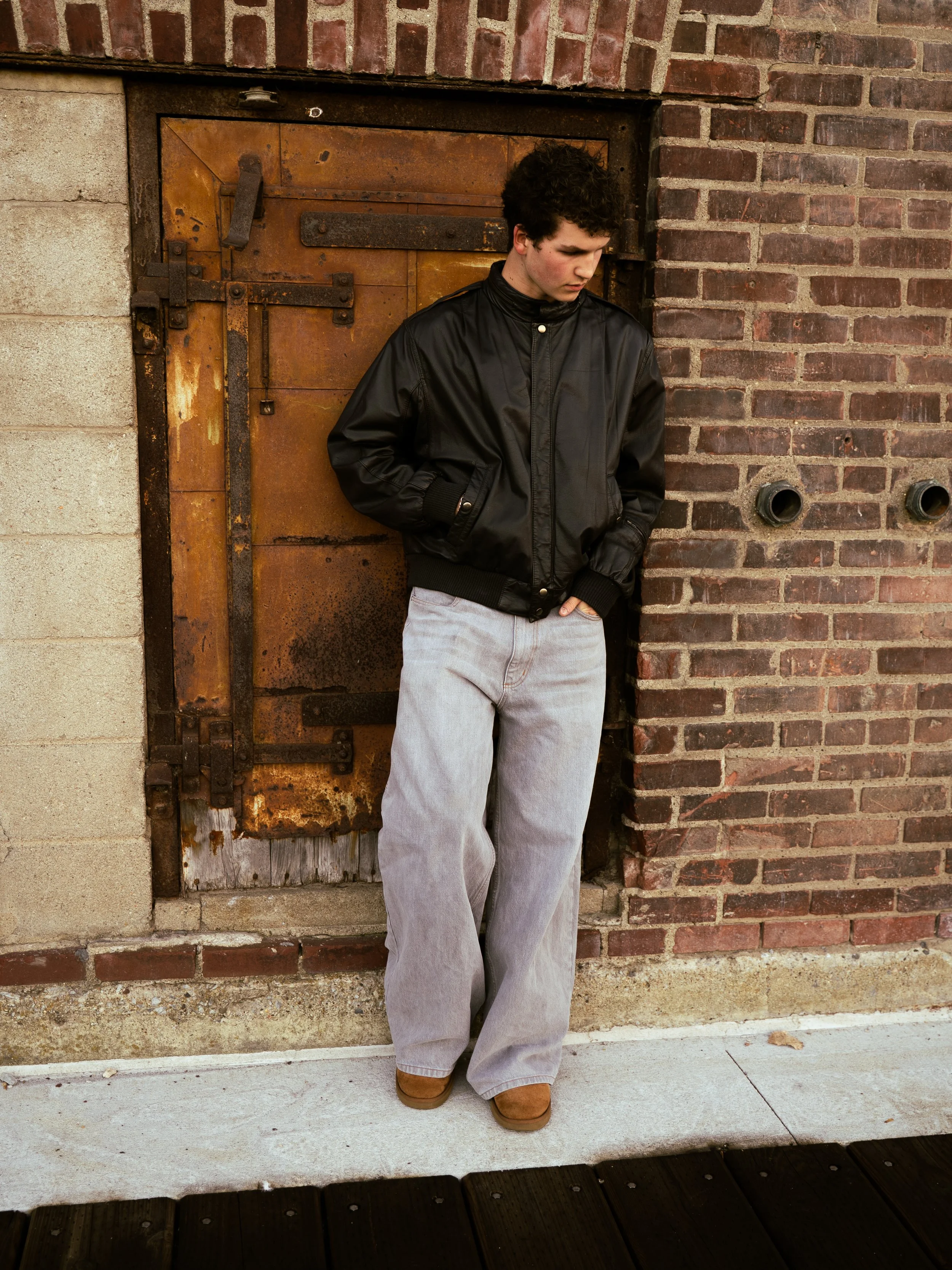 A young man with curly dark hair wearing a black leather jacket and light gray pants standing against an aged brick and concrete wall near an old rusty metal door.
