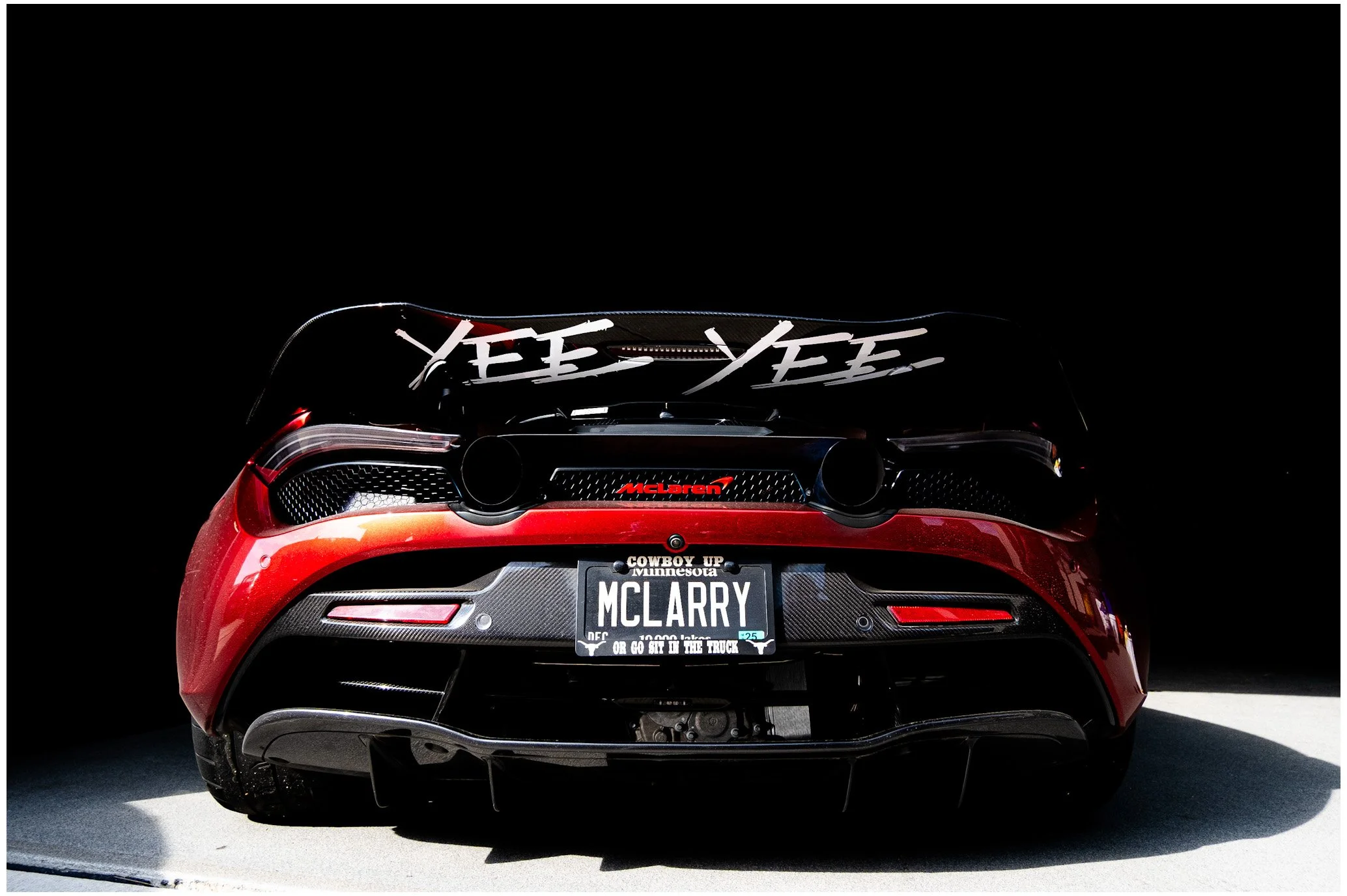 The rear view of a red McLaren sports car with custom license plate that reads "McLARRY". The car has a black spoiler with the words "YEE YEE" written in white on it. The background is dark, making the car the focal point of the image.