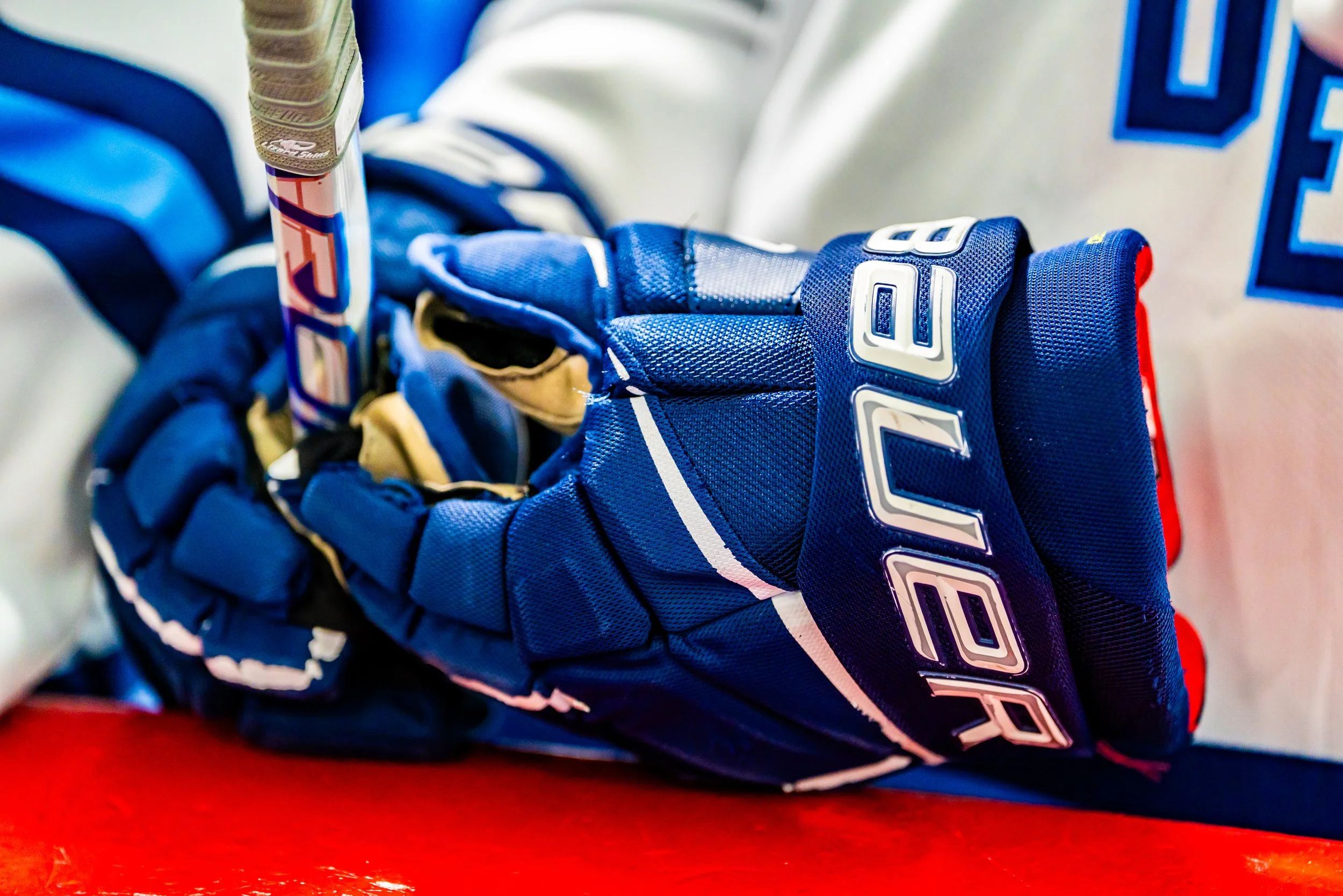 Close-up of a blue hockey glove with white and black accents resting on a red surface. Part of a hockey stick is visible, with the blade partially covered by the glove.