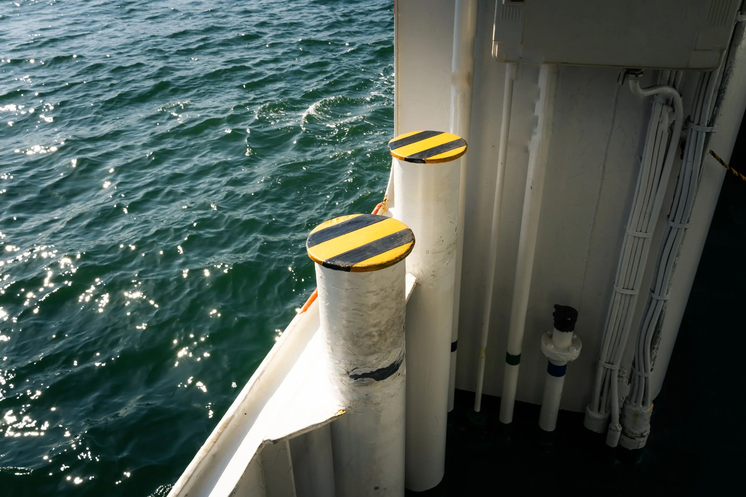 View of calm ocean water from a boat, with part of the boat's side visible including four vertical safety posts with yellow and black striped tops, and some piping and cables.