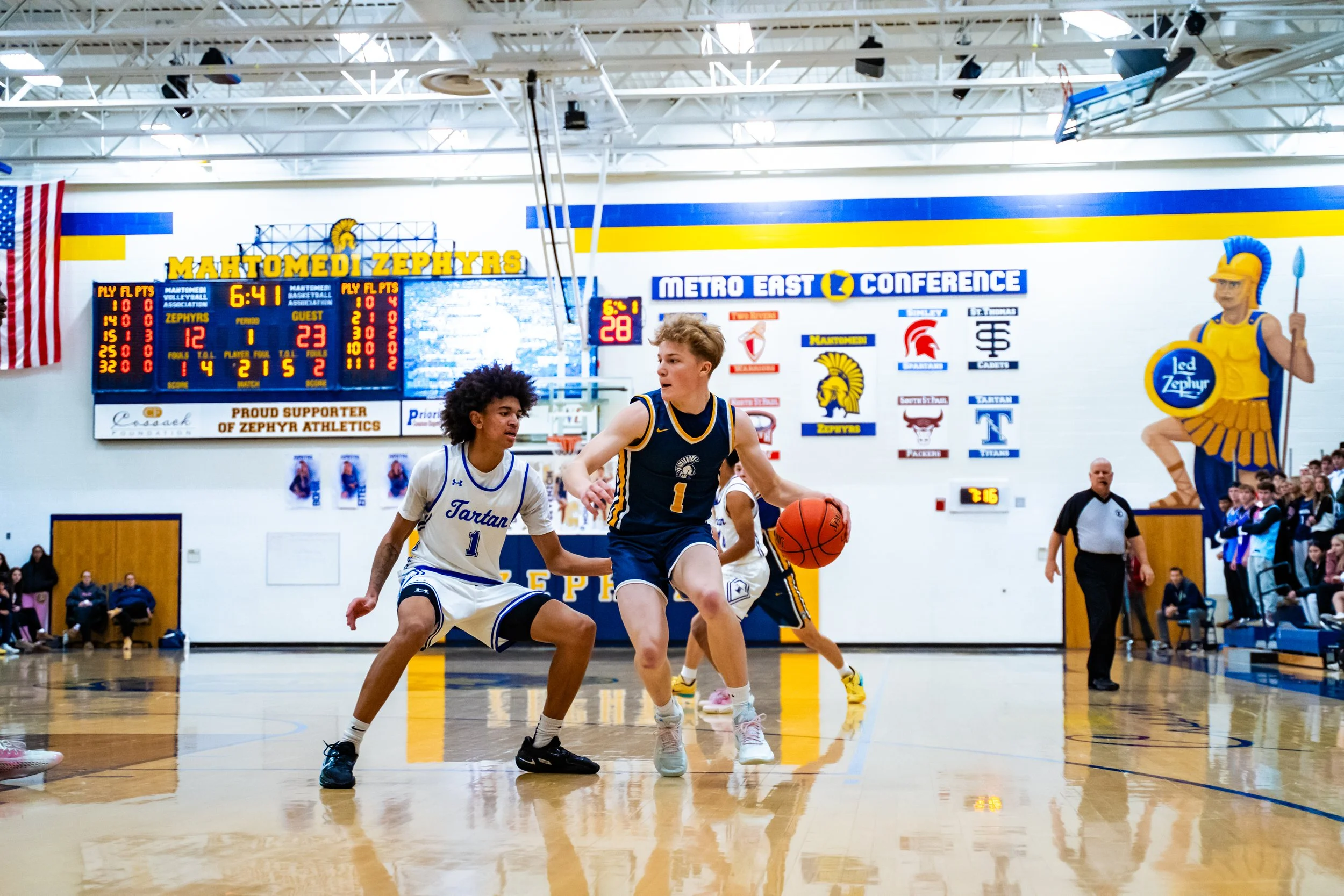 Two young basketball players compete for the ball during a game in an indoor gymnasium, with spectators and a referee in the background.