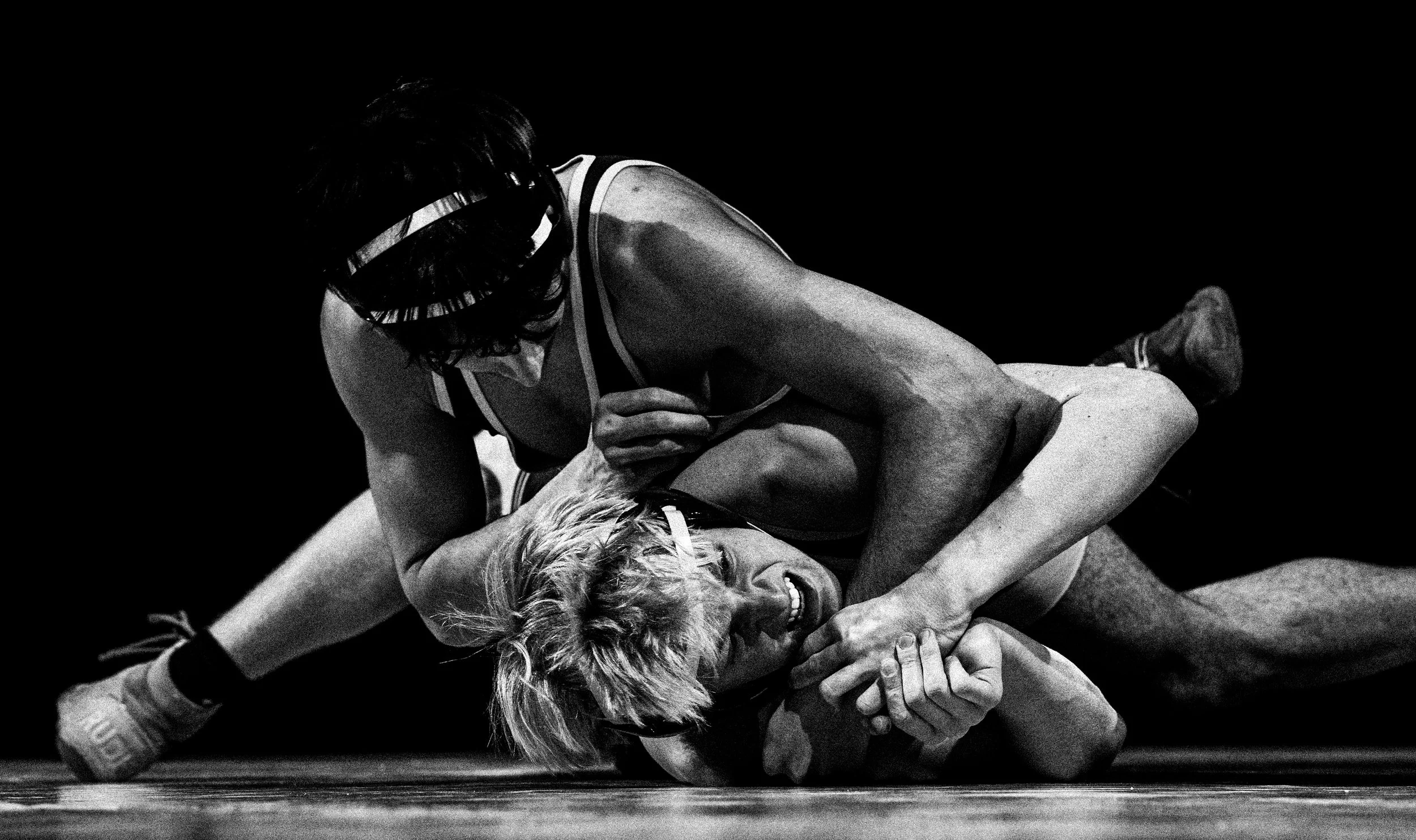 Two women wrestling on a mat, one woman with blonde hair and one with dark hair, both wearing athletic clothing. The woman with dark hair is on top, gripping the blonde woman's arm and shoulder, with a focused expression.