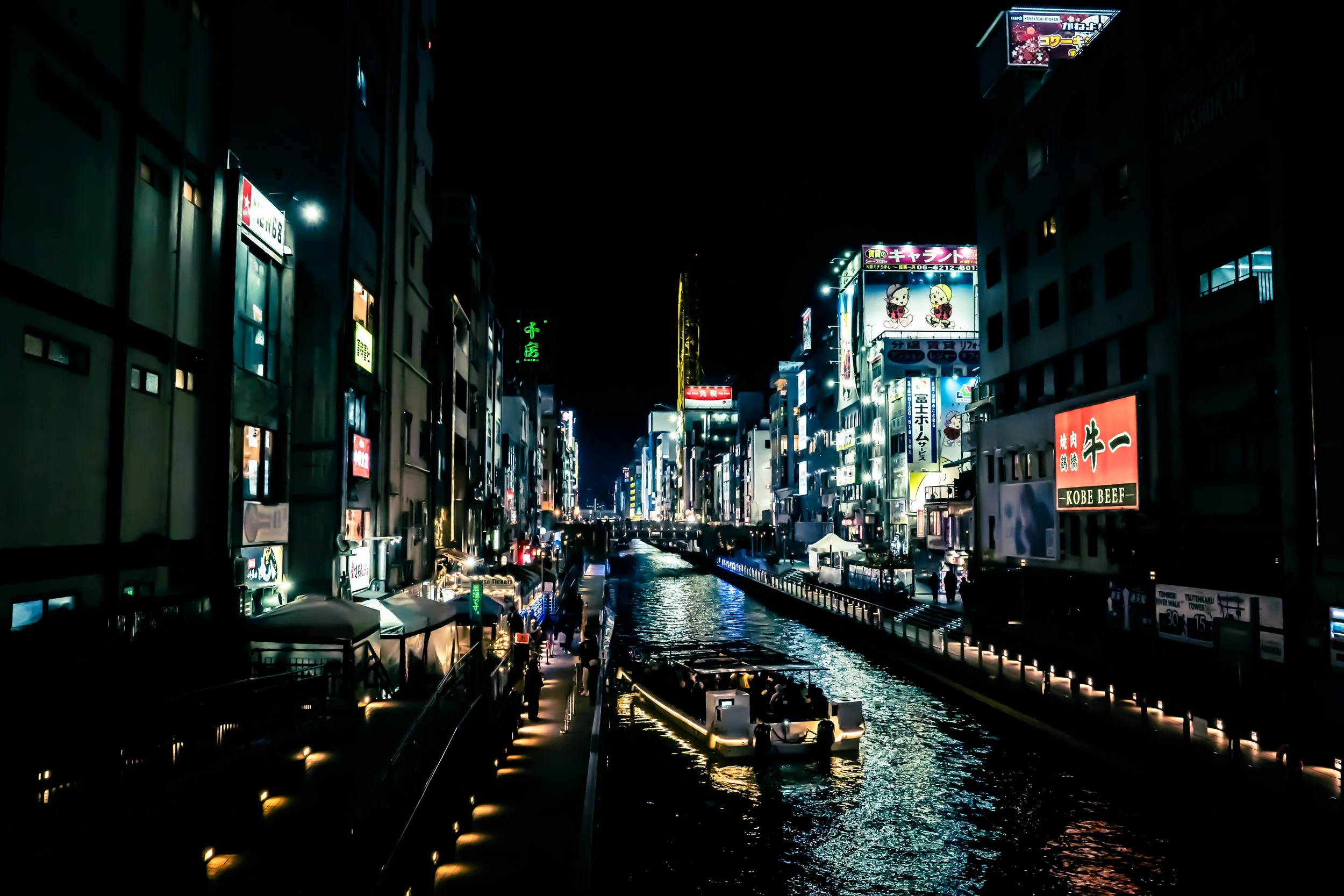 Nighttime scene of a busy city street with illuminated signs, buildings, and boats on a canal, likely in Japan.