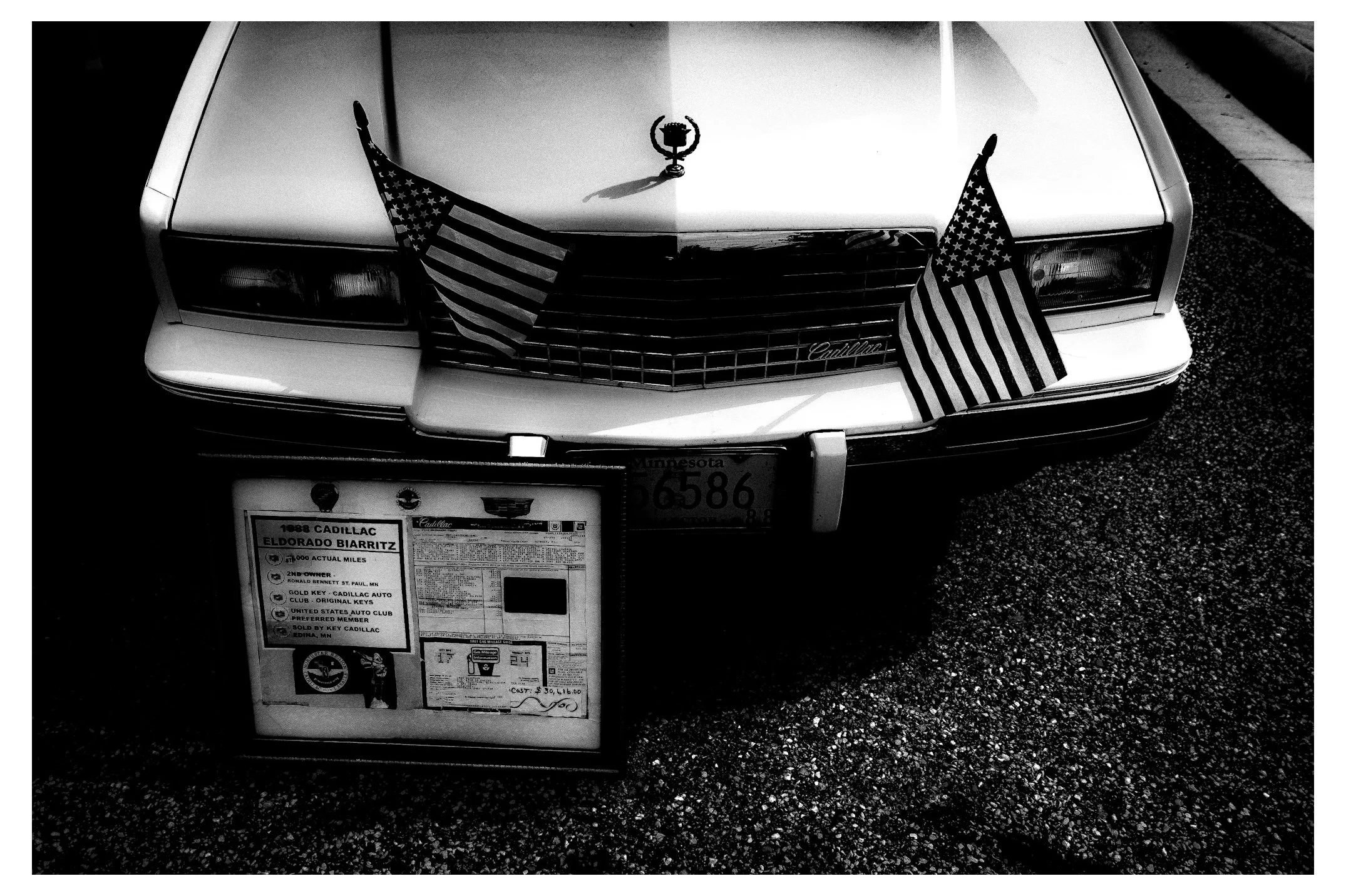 Front view of a classic white Cadillac car with two American flags attached to the front grill. The car has a hood ornament and a license plate from Minnesota. A large informational window sticker is displayed on a stand in front of the car.