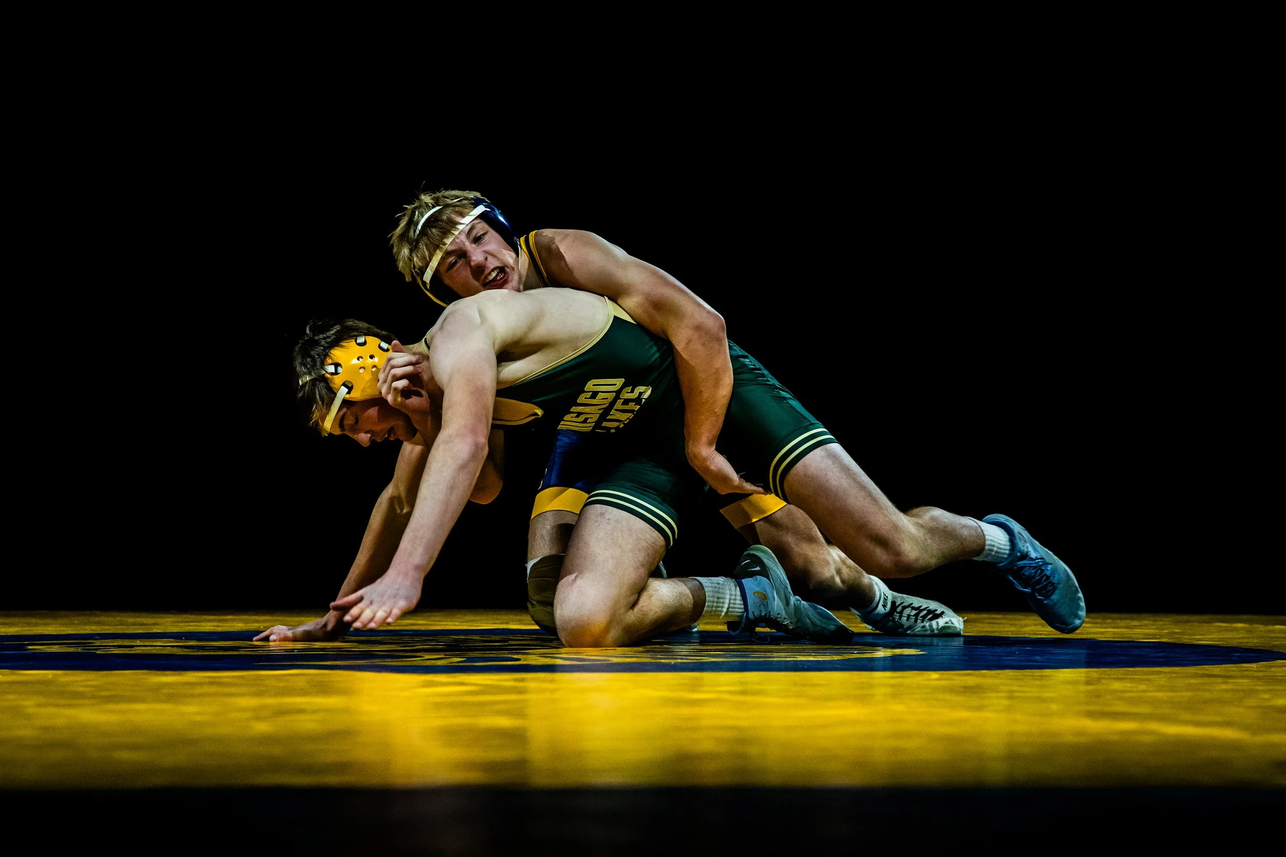 Two high school wrestlers compete on a yellow wrestling mat; one in black and gold singlet and the other in dark green singlet, with a black background.
