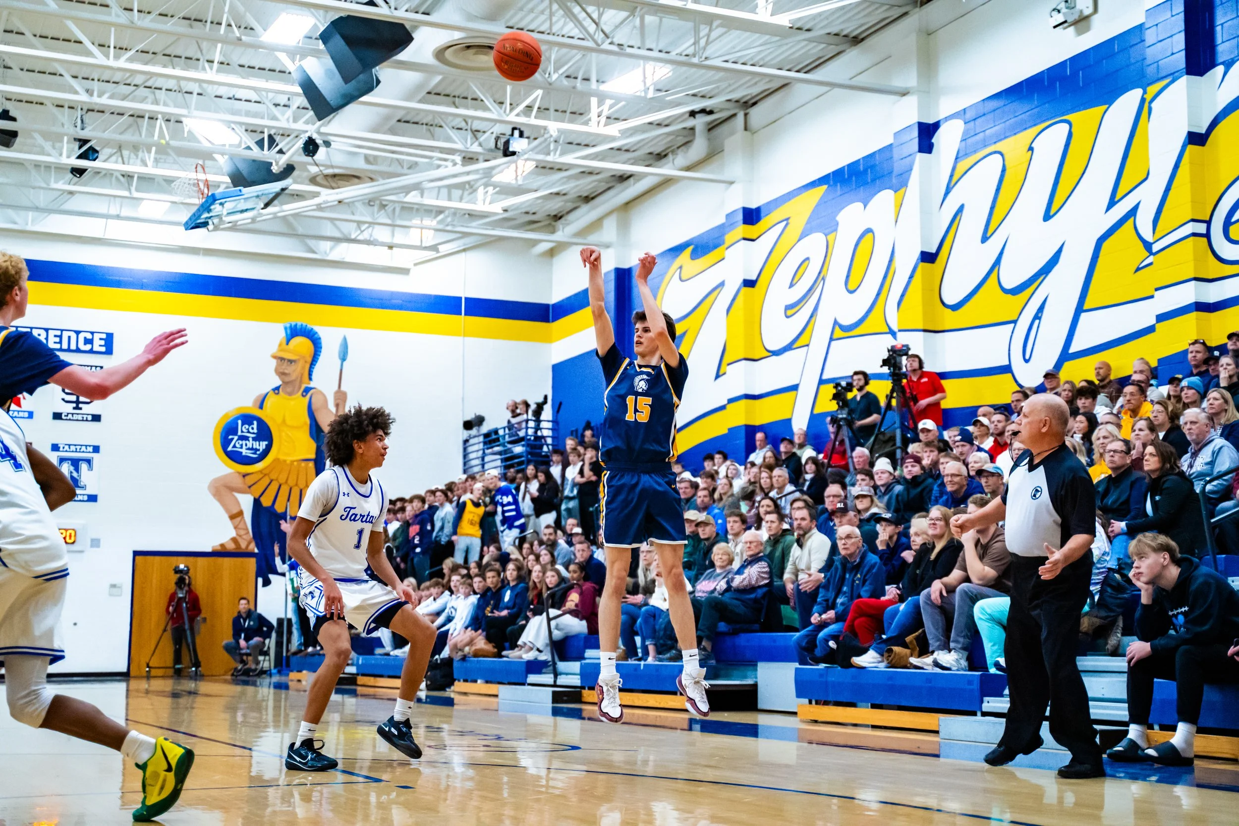 A basketball game taking place in a gymnasium filled with spectators. A player in a blue uniform, number 15, is jumping to shoot the ball while another player in white tries to block. The gym has a large wall with the word 'Troy' painted in yellow an