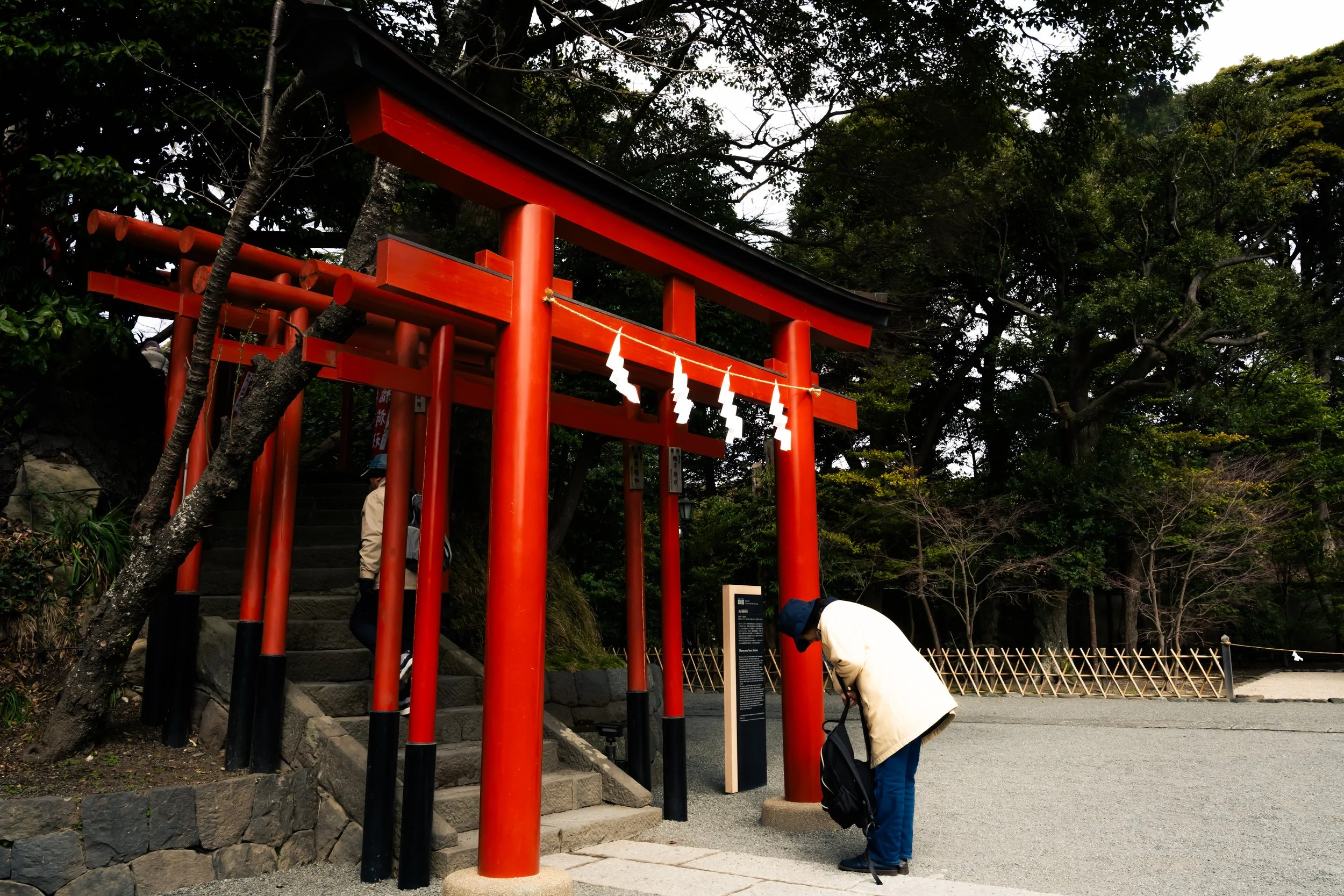 A red torii gate at a shrine entrance surrounded by trees, with a person in a white coat and blue pants leaning over near the gate.