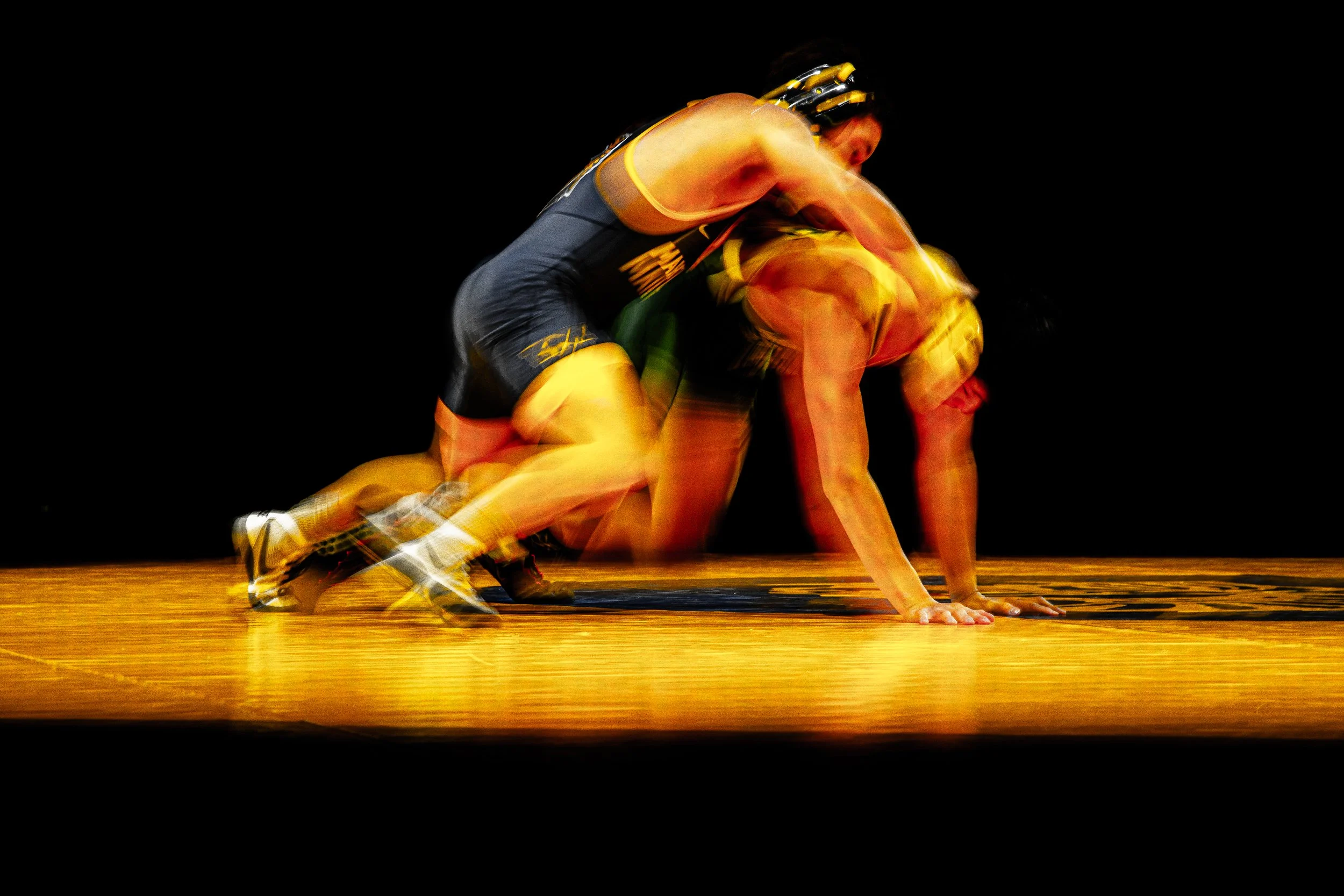 Two athletes in athletic gear perform a wrestling move on a wooden competition mat, with one person on top of the other, against a black background.