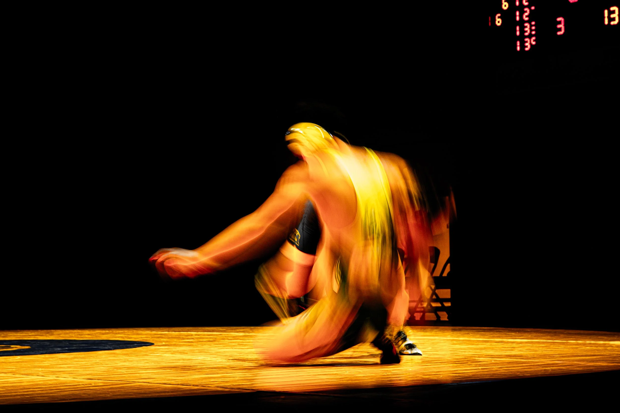 Blurred image of a person in yellow sportswear practicing on a wooden floor, with a digital clock/display in the background showing red numbers.