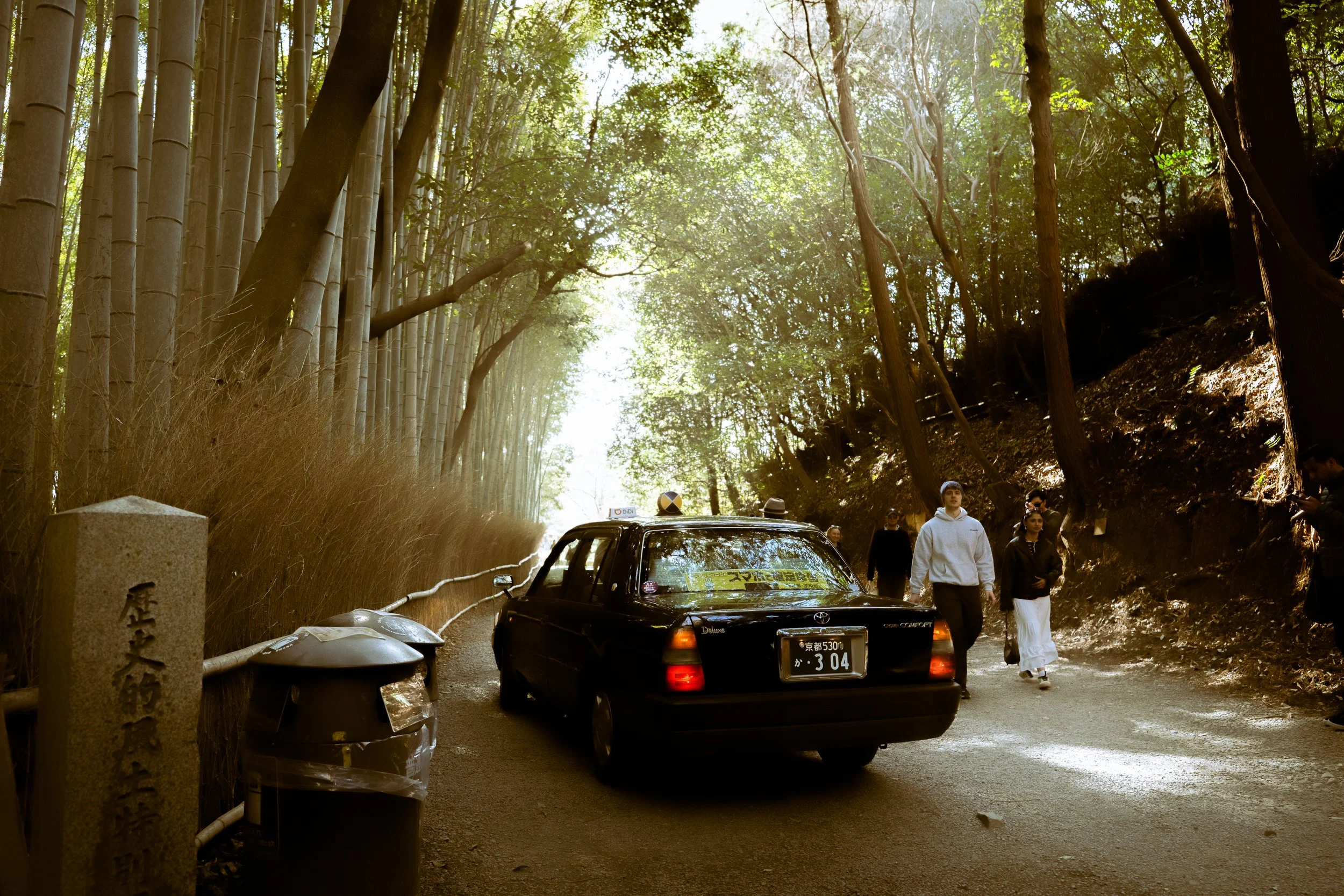People walking along a path in a forested area with tall trees. A black car is parked nearby, and there are trash bins and stone markers along the path. The sunlight filters through the leaves, creating a dappled light effect.