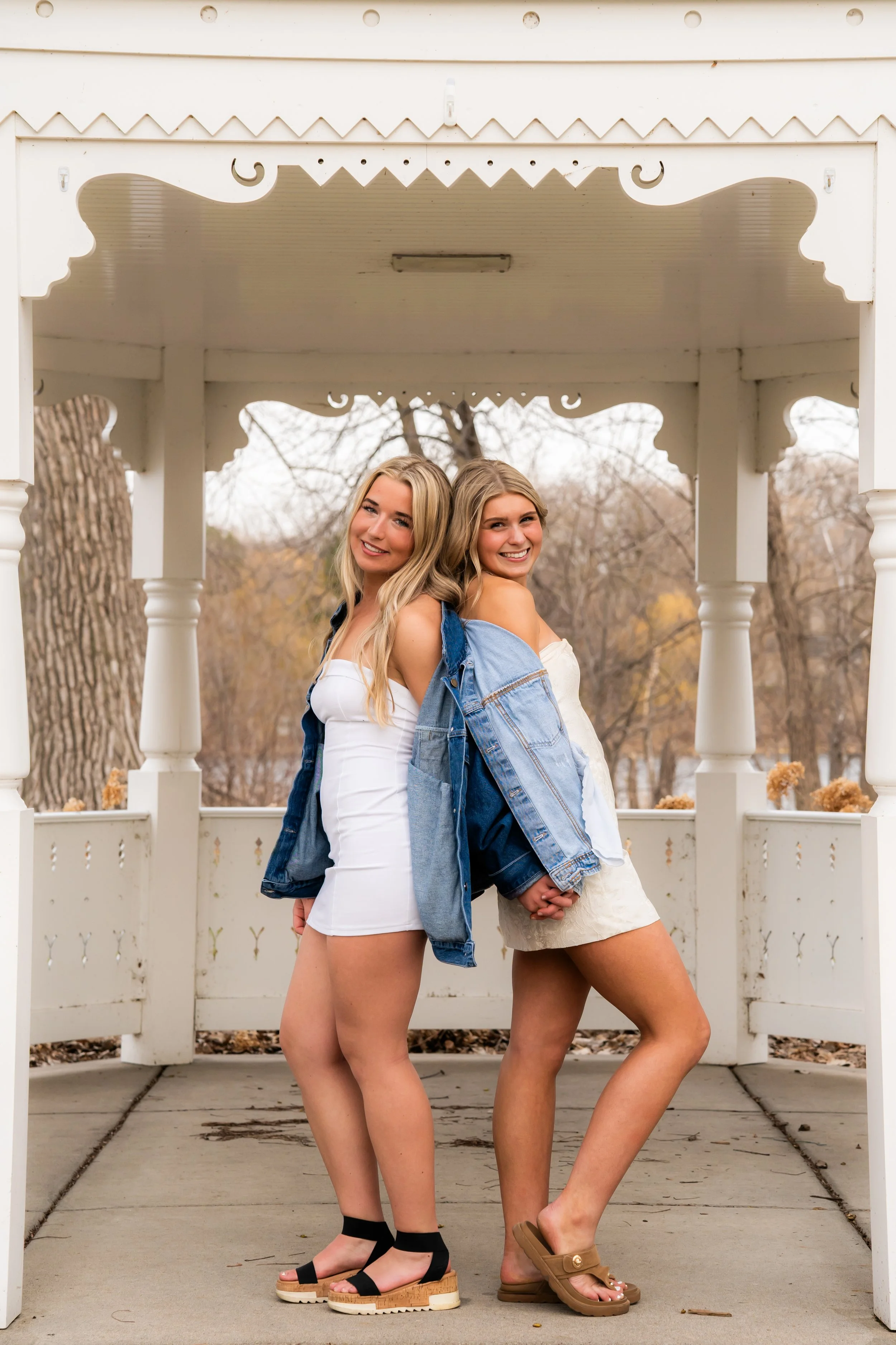 Two young women standing back to back under a decorative white gazebo in an outdoor park, holding hands and smiling.