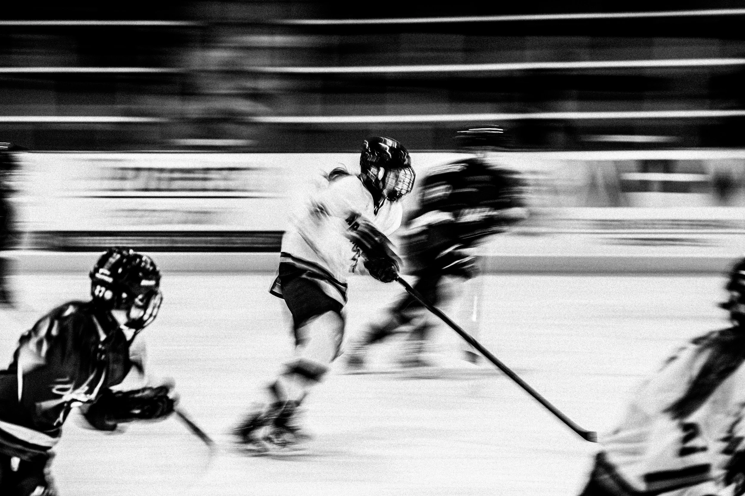 Black and white action shot of a hockey game with players in motion, including a goalie on the ice and players with hockey sticks.