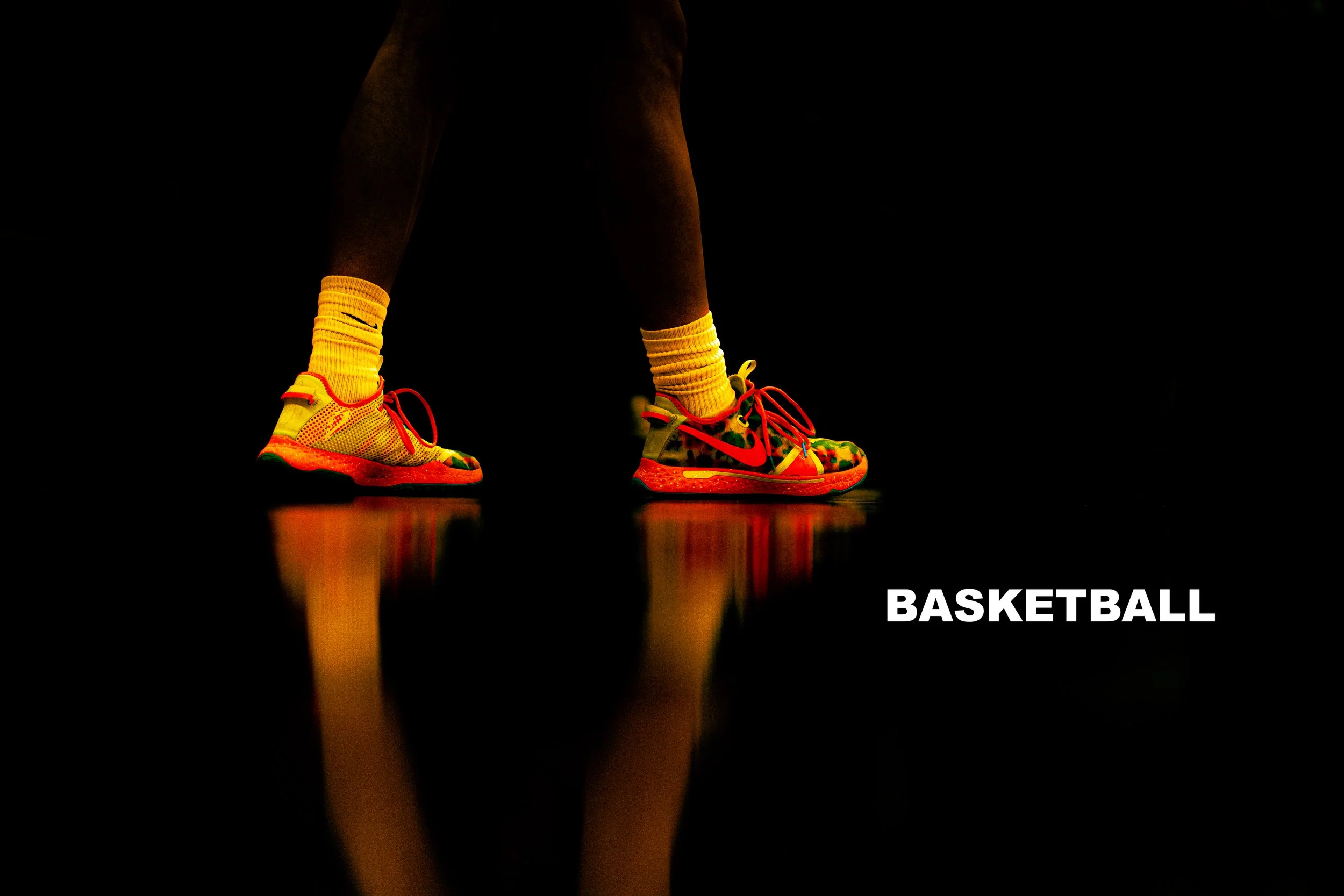Close-up of a person's legs wearing colorful basketball shoes and yellow socks standing on a reflective black surface with the word 'BASKETBALL' written in white at the bottom right corner.