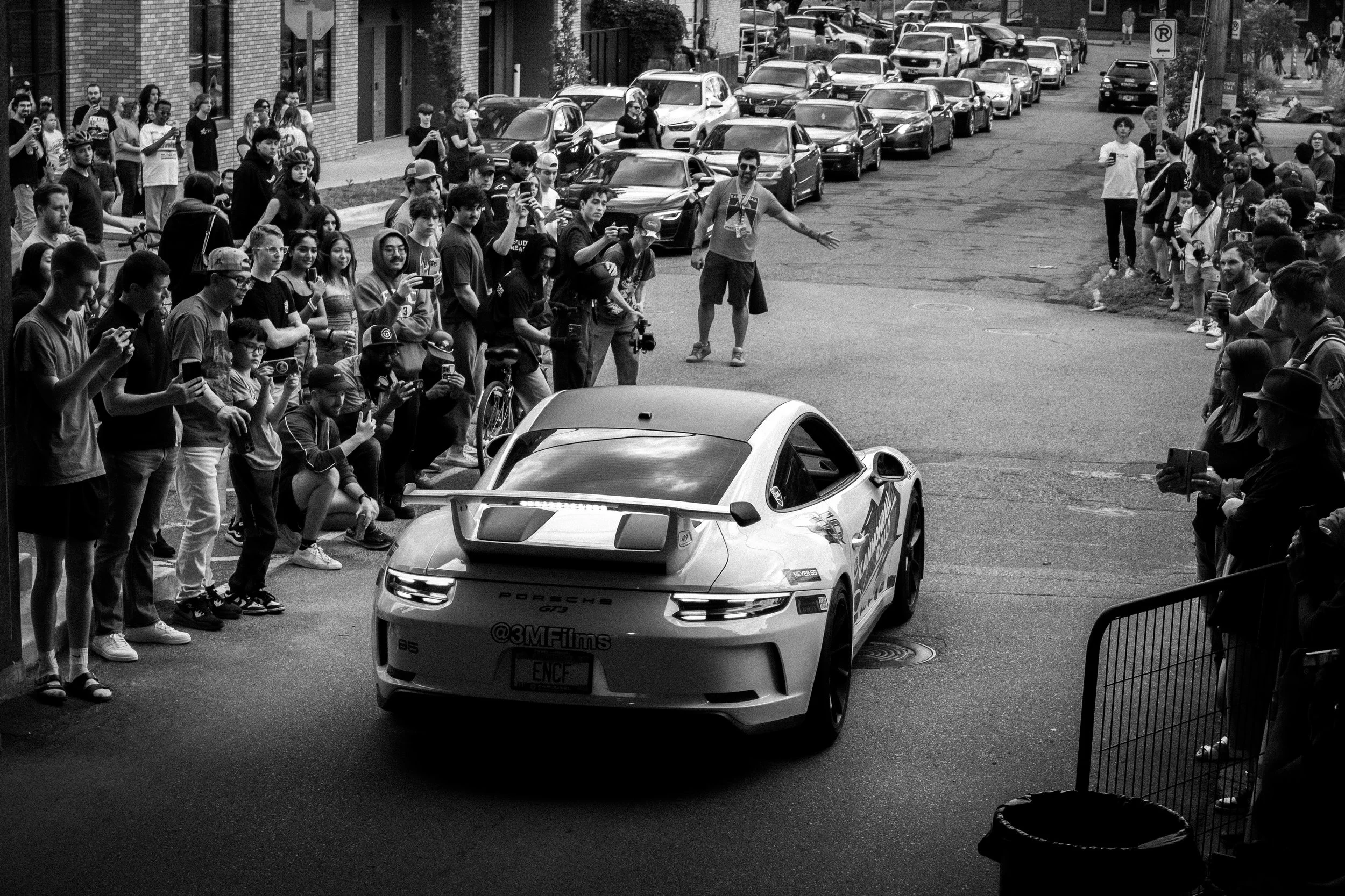 A crowd of people gathered on a city sidewalk and street to watch a race car passing by, with many taking photos or videos, with parked cars and buildings in the background.