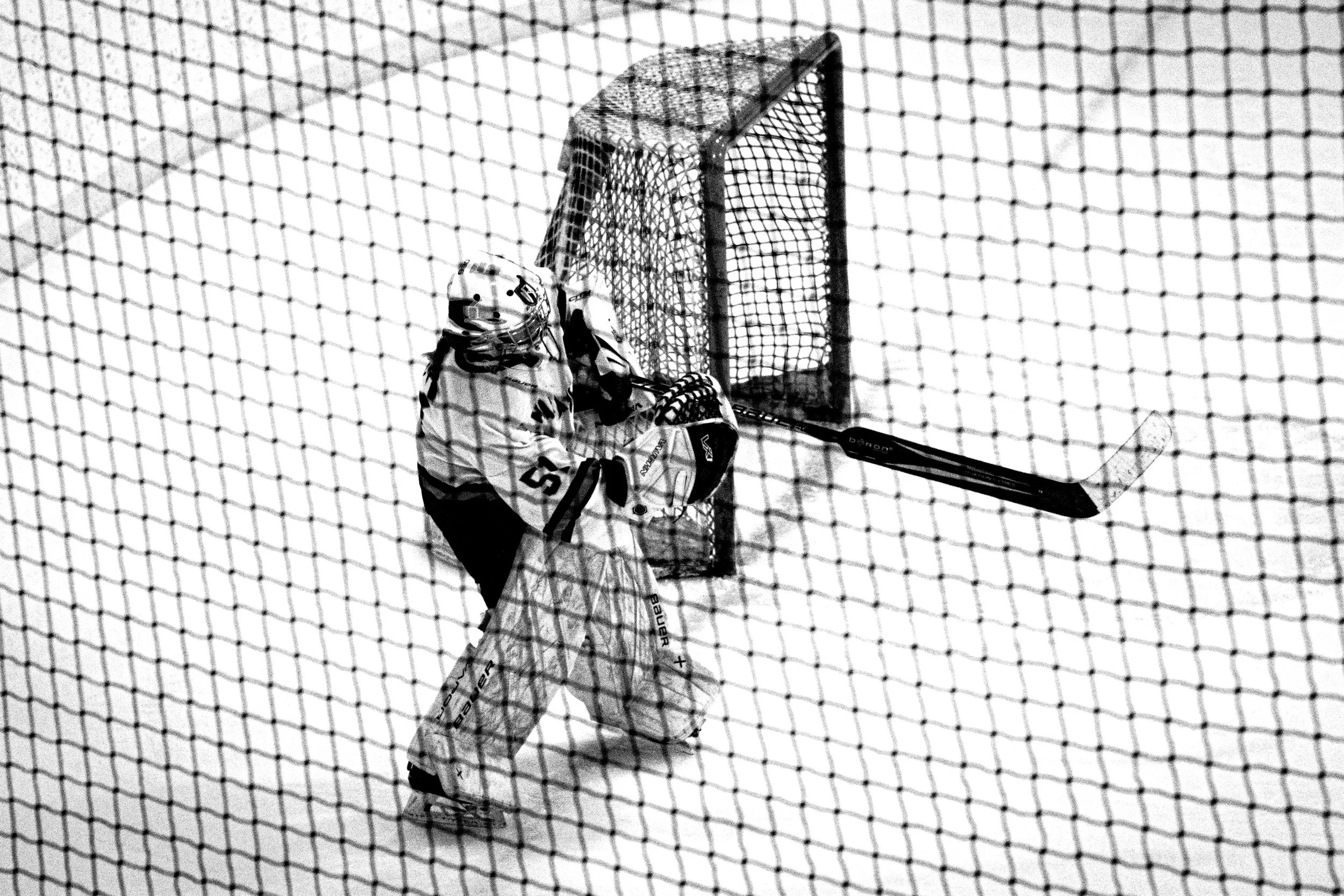 A hockey goalie in a uniform and helmet stands in front of the net on an ice hockey rink, seen through the protective netting.