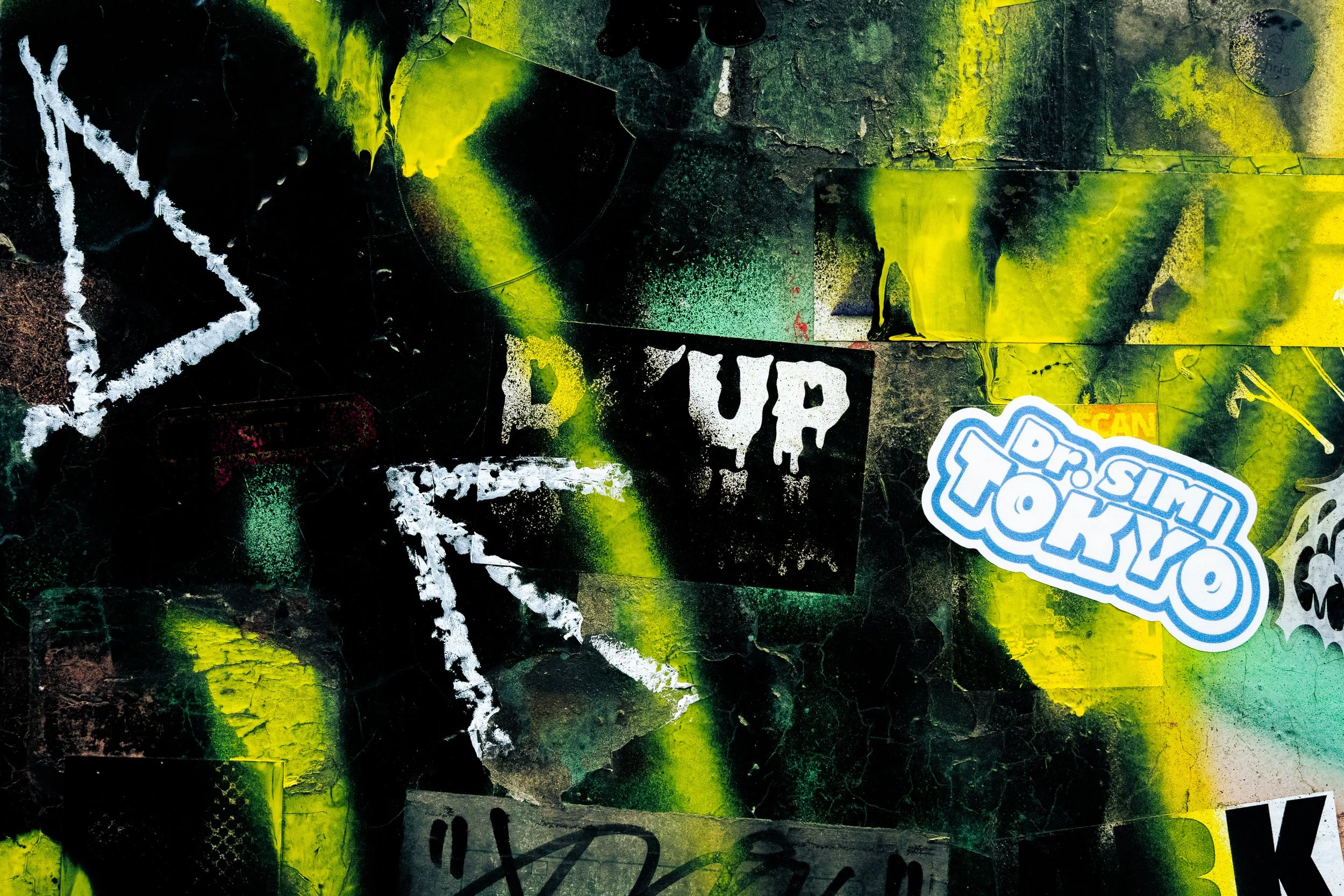 Close-up image of a vibrant graffiti-covered wall with splashes of neon yellow, black, white, and green paint, featuring various stickers and street art tags.