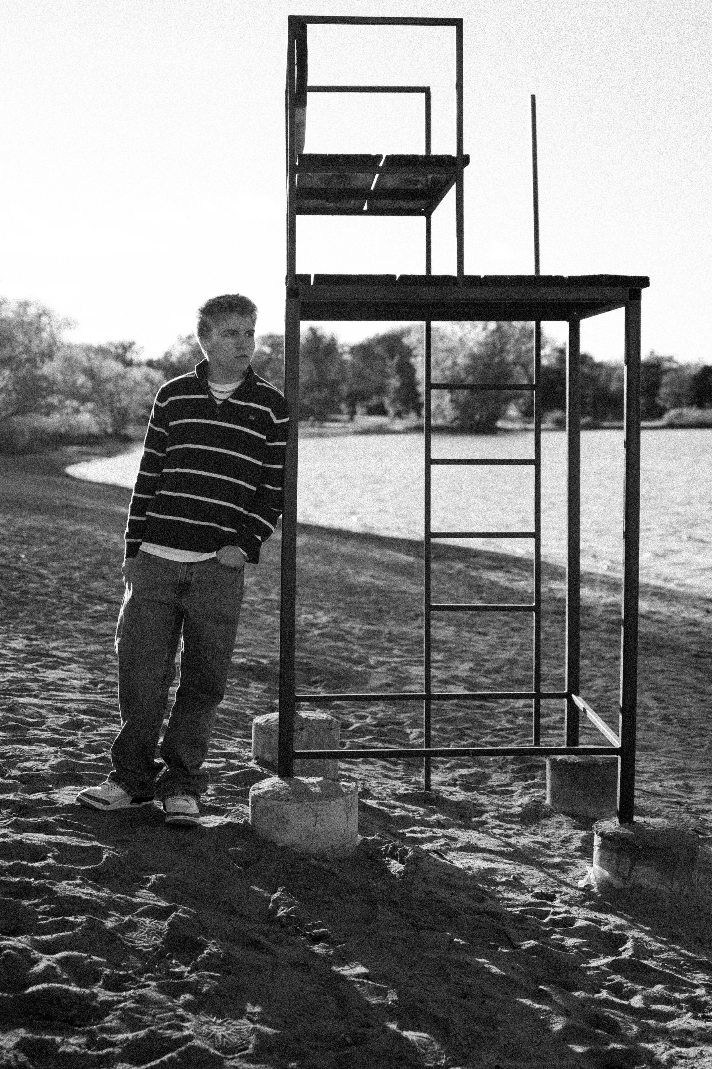 Black and white photo of a young boy standing on a sandy beach near a metal structure with several levels, possibly a lifeguard stand. The boy is wearing a striped jacket, jeans, and sneakers, and is looking to the side. A body of water and trees are
