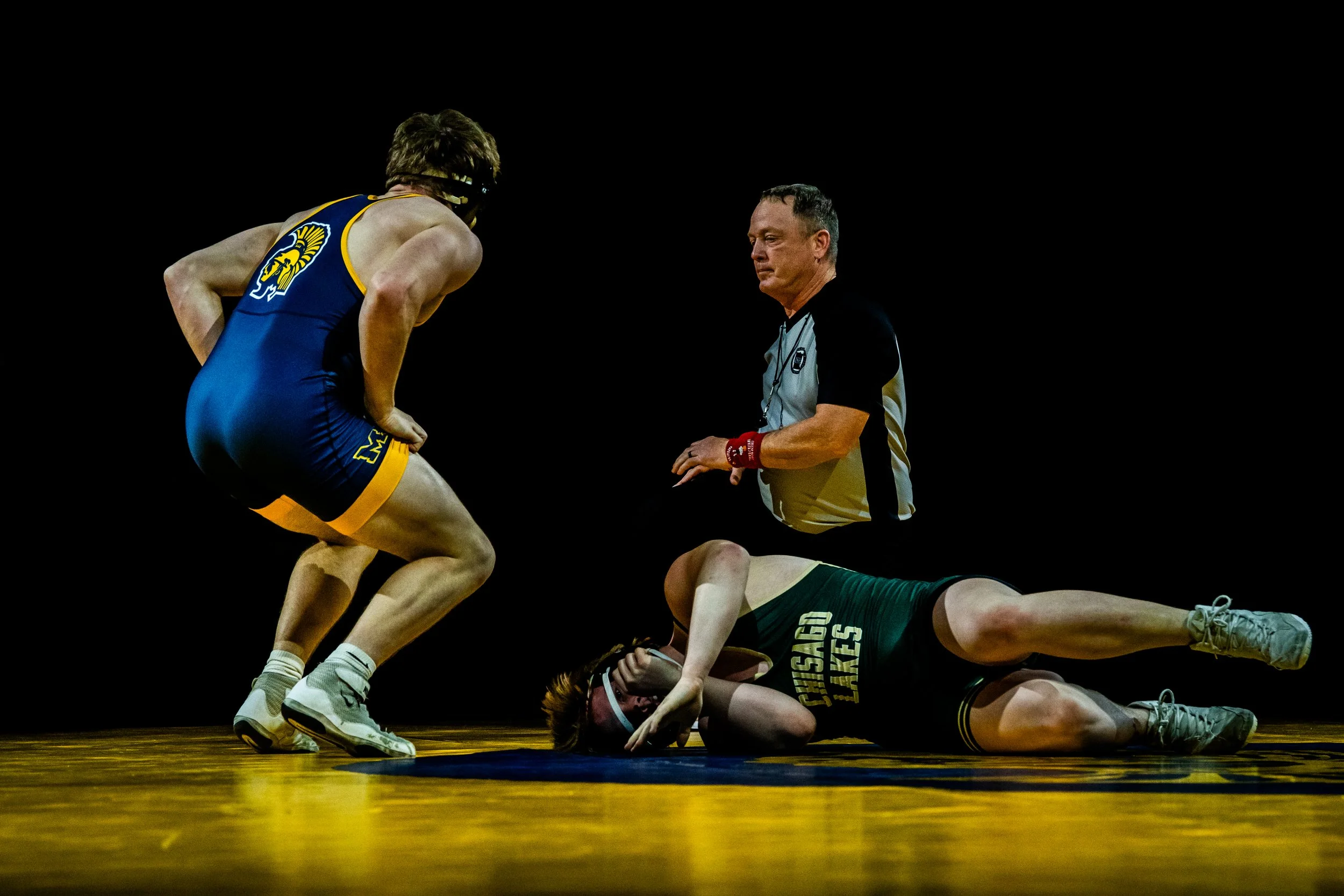 Wrestler in blue singlet standing over opponent on the mat, with referee observing, during a wrestling match.