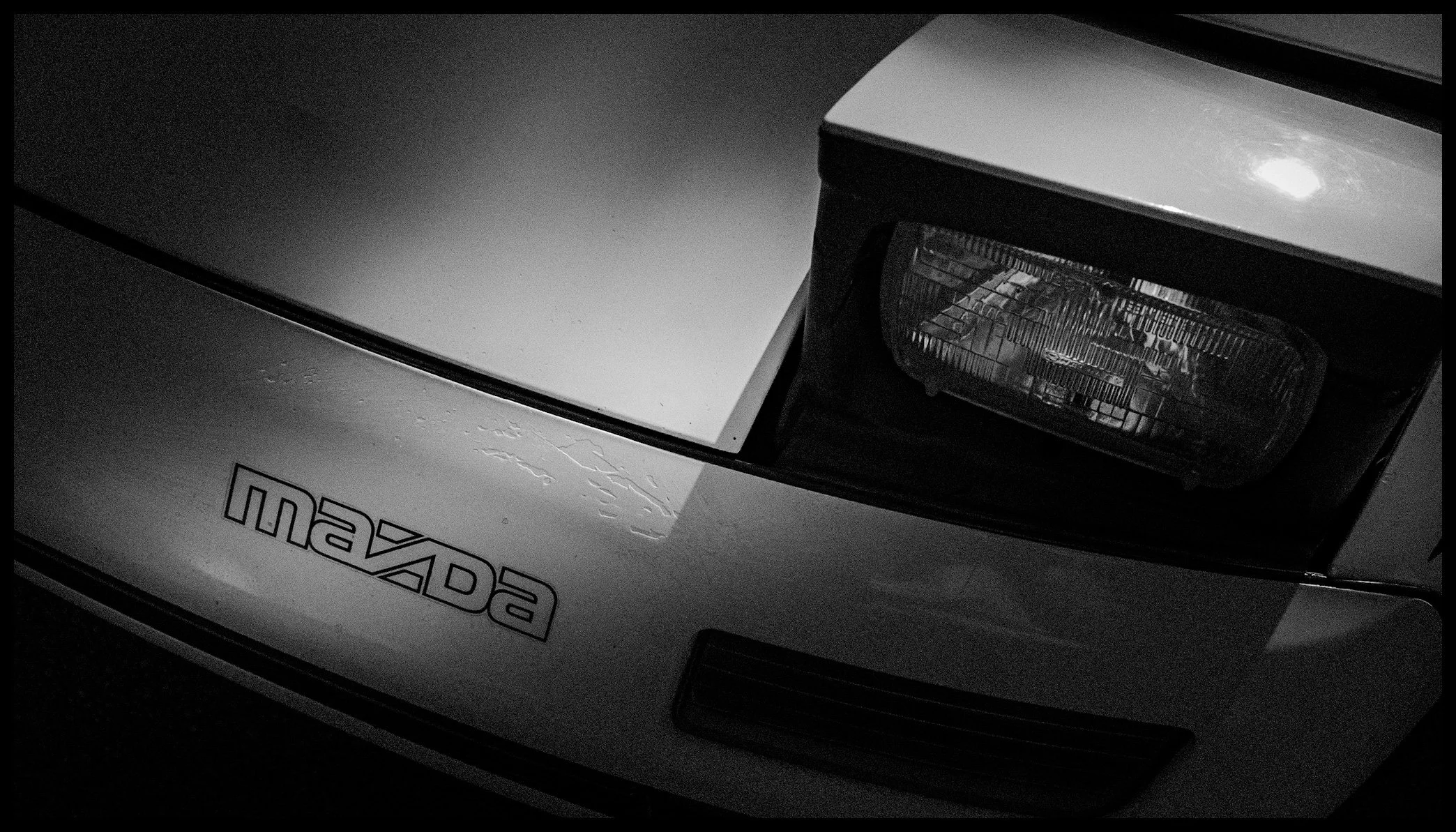 Close-up of a MaXcar vehicle, showing part of the hood and front light in black and white.