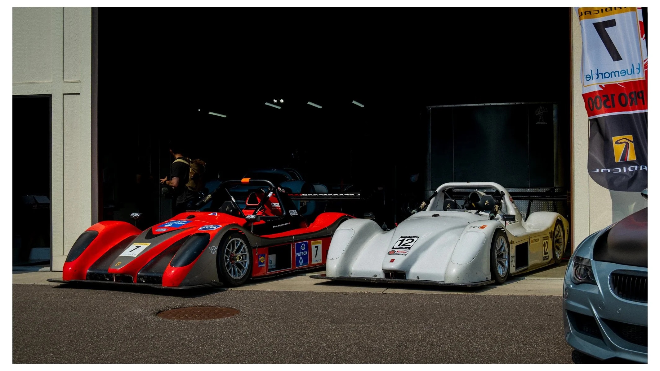 Two race cars, one red with black accents and one white, parked side by side outside a building, with a person in the background near the entrance.