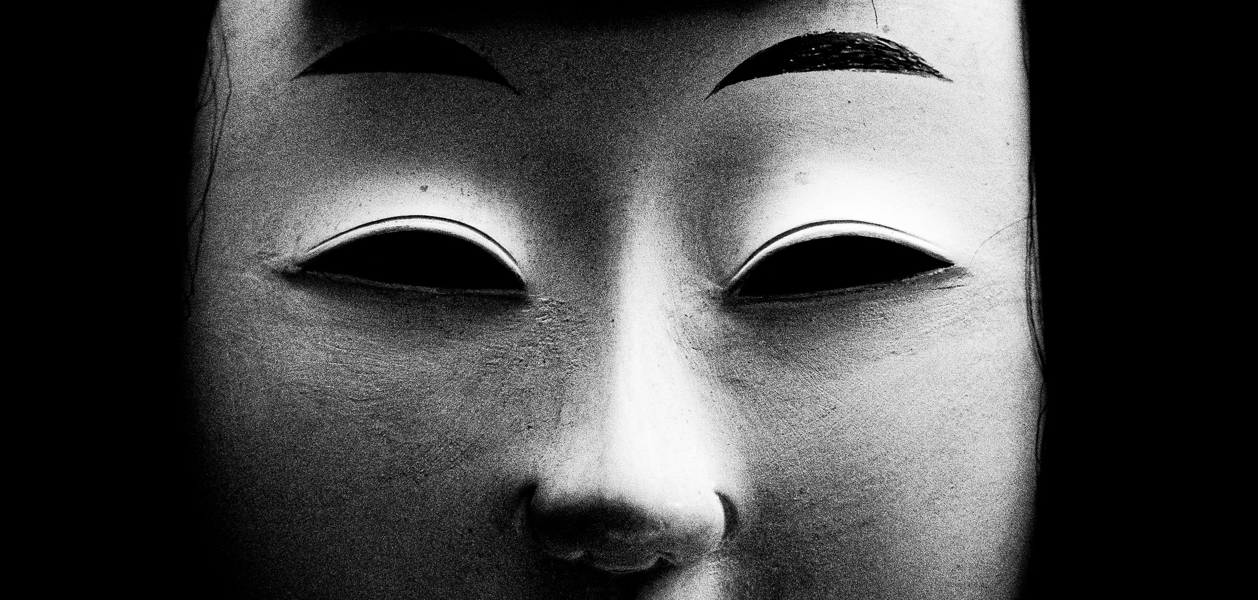 Close-up of a traditional Asian mask with a smooth, light-colored face, dark eye openings, and dark eyebrows set against a black background.