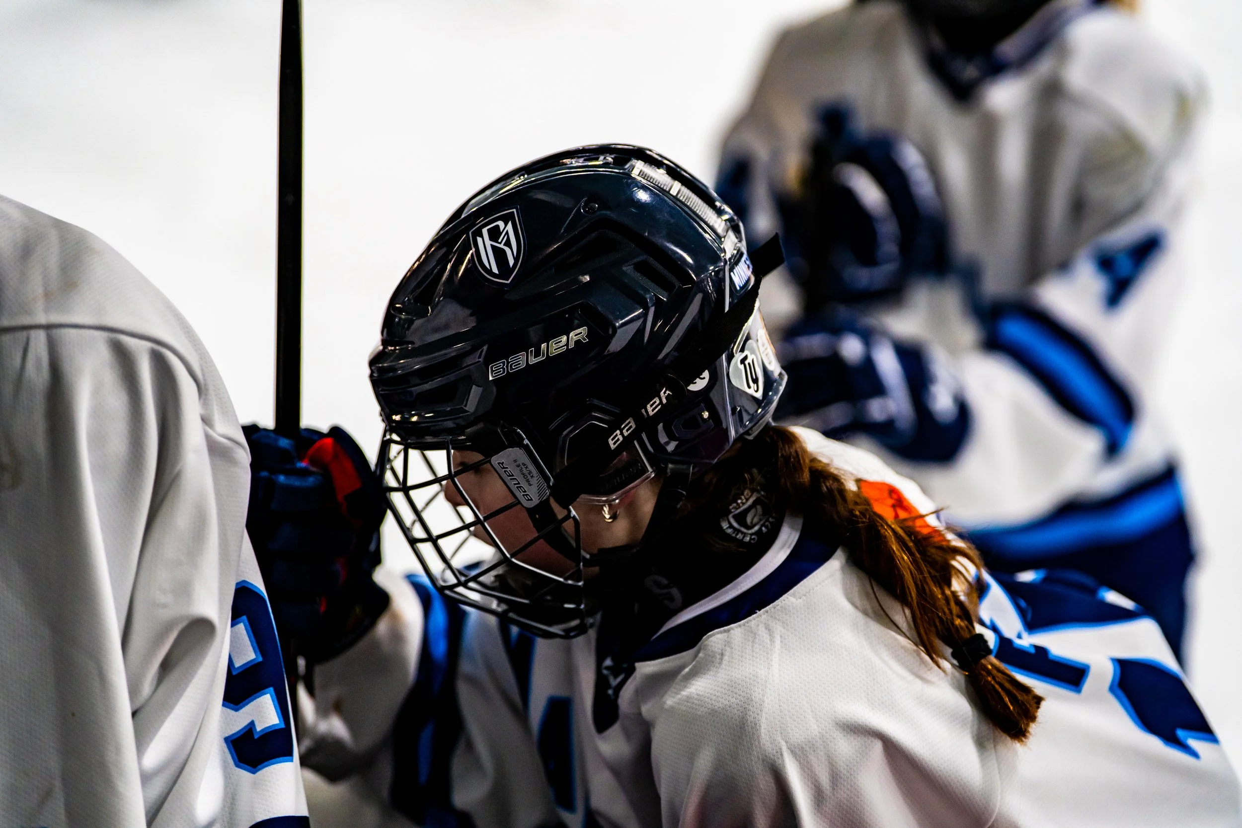 Close-up of a female ice hockey player wearing a black Bauer helmet, with teammates in white jerseys around her.
