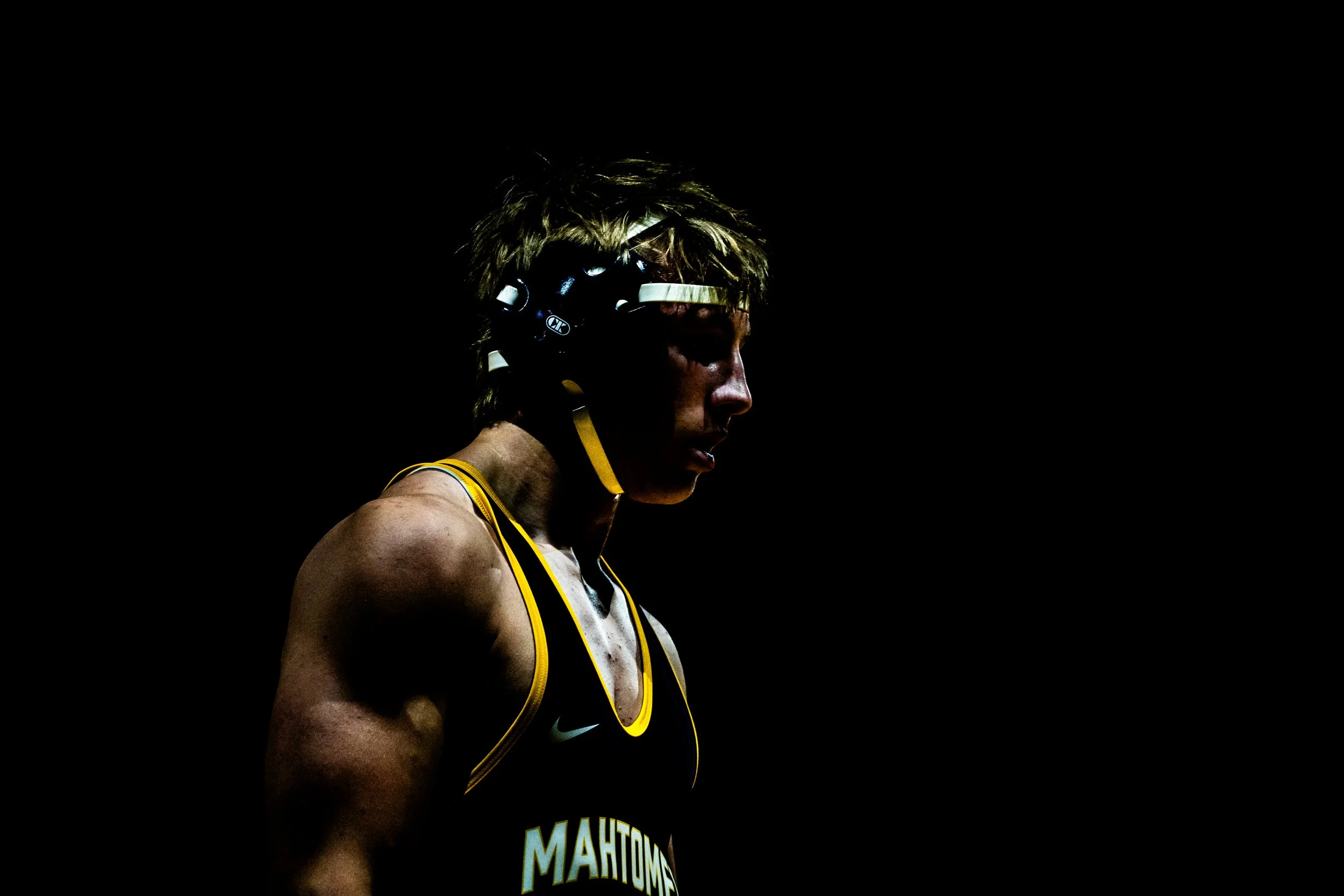 A female athlete with short hair wearing a black and yellow sports uniform and a helmet, standing in profile against a dark background.