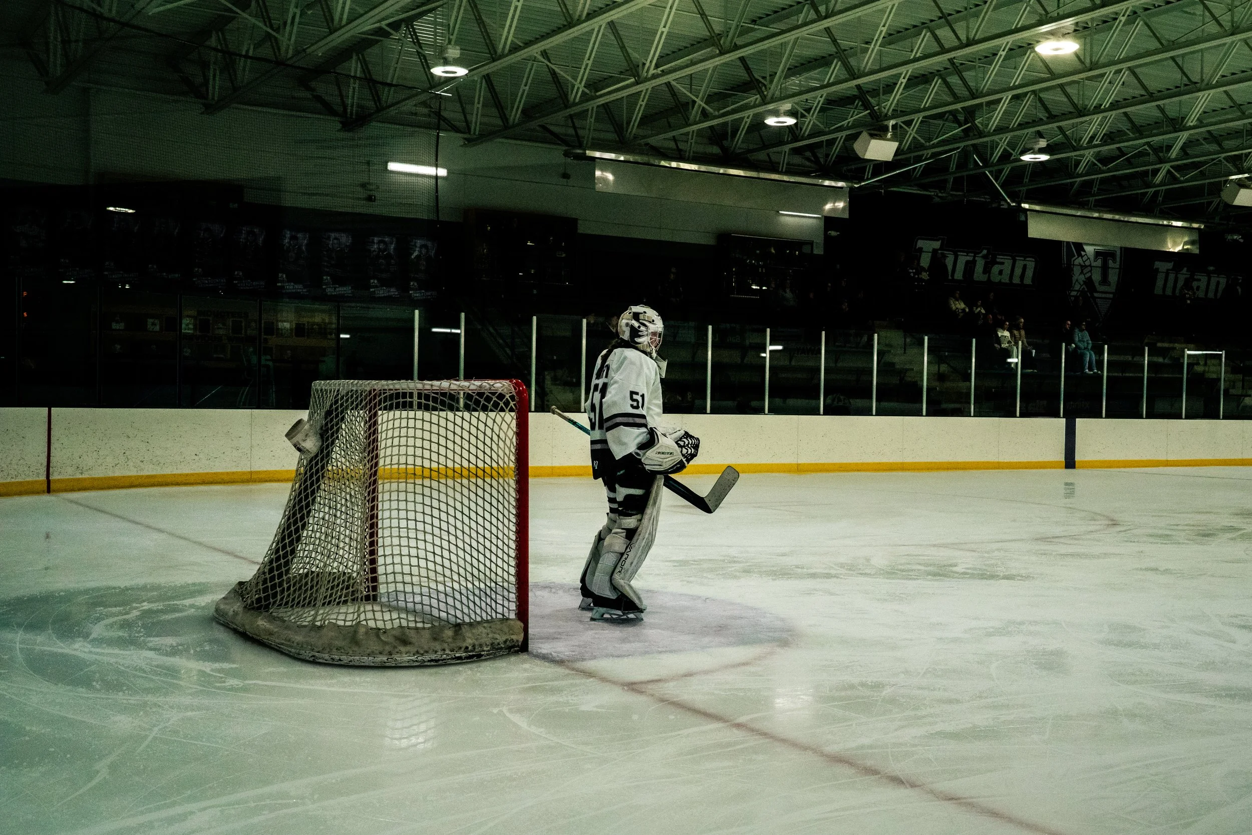 An ice hockey rink with a goalkeeper in white and black gear standing near the goalpost. The rink is enclosed with a high glass barrier, and the background shows seating and the arena interior with high ceiling lights.