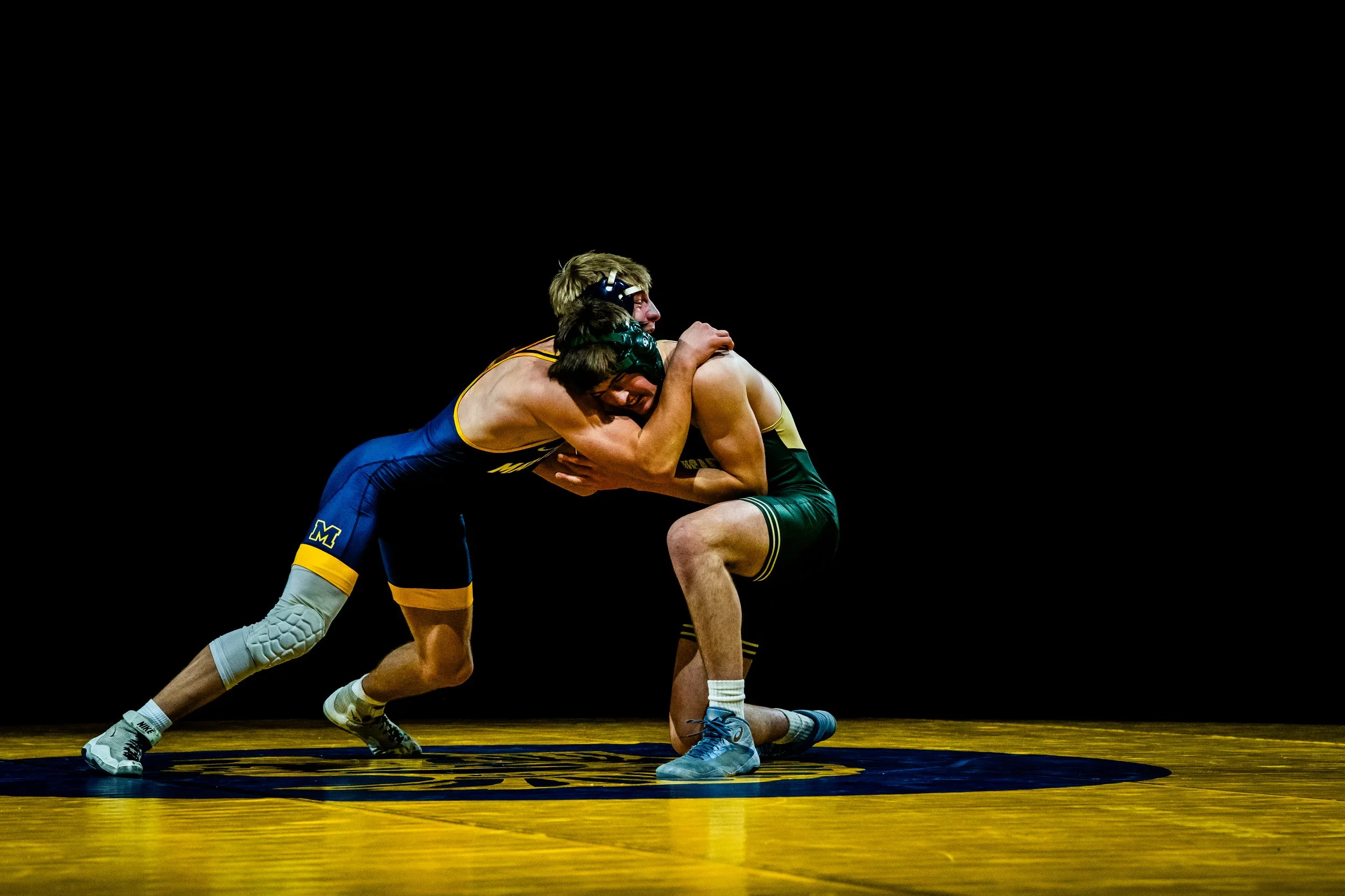 Two male wrestlers engaged in a match, one in a blue and yellow singlet and the other in a black and yellow singlet, wrestling on a yellow mat with a black logo, under black lighting.