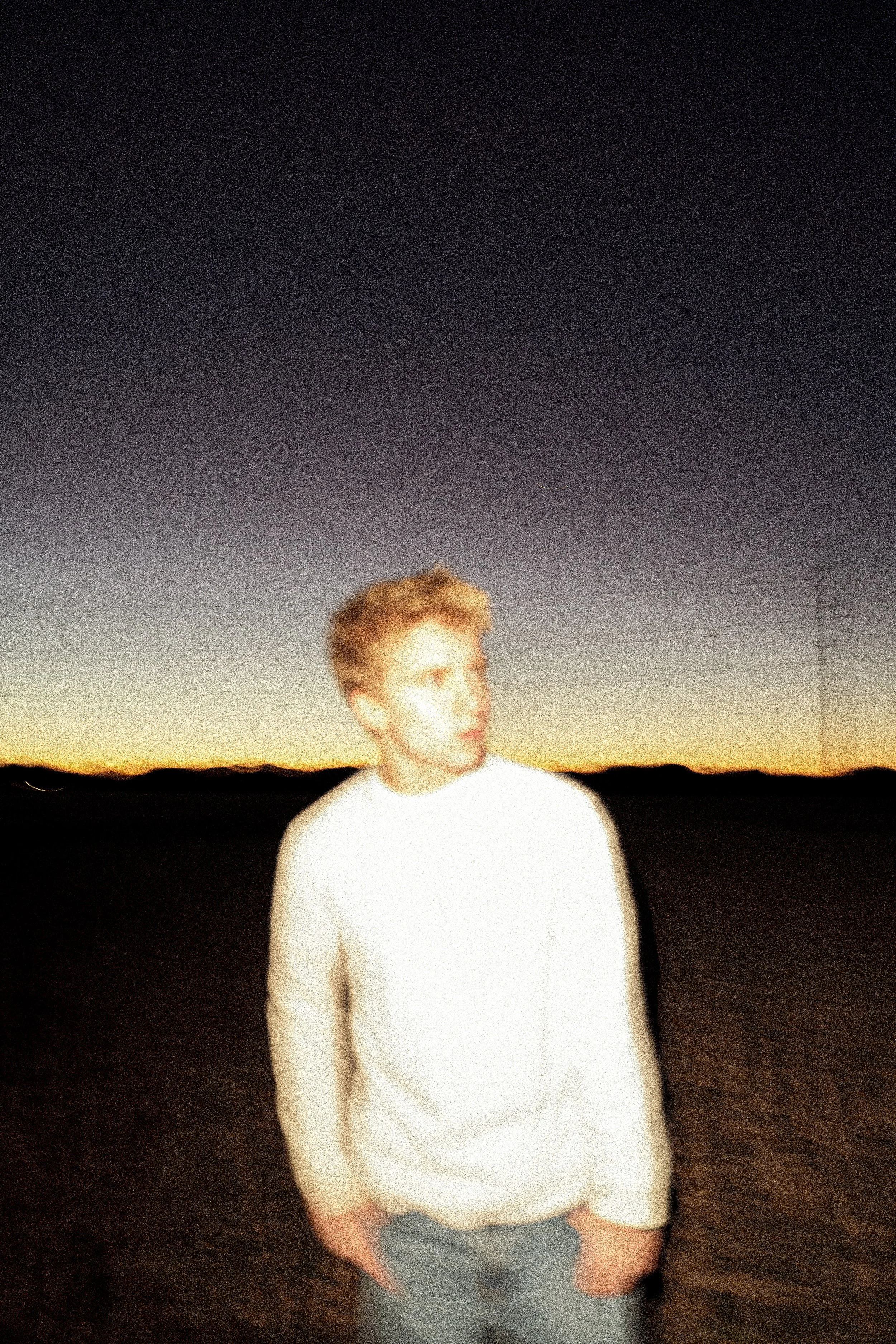 A person standing outdoors at dusk or night, with a starry sky and a distant horizon in the background, wearing a white shirt and jeans.